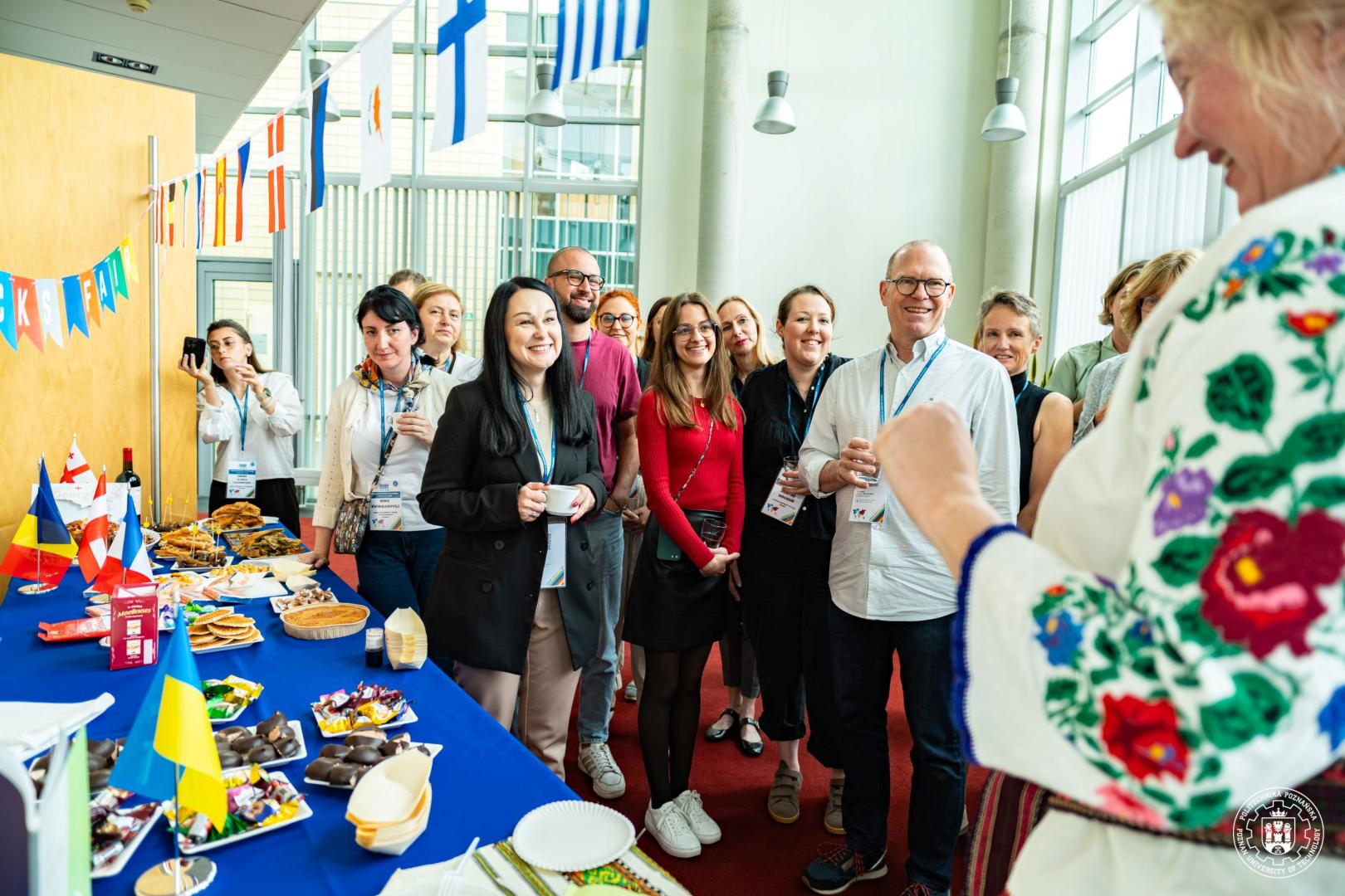 Participants gathered around a table with regional snacks and national flags, listening to a woman in a traditional Ukrainian embroidered shirt