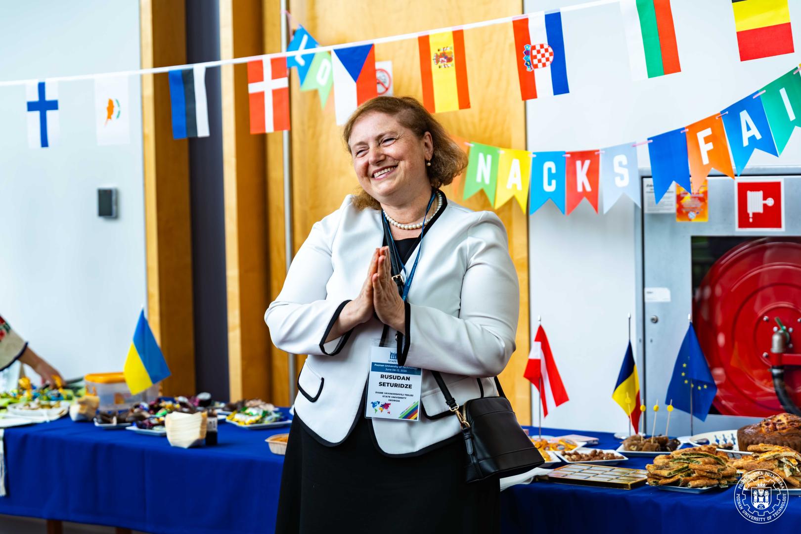 A smiling Rusudan Seturidze with her hands folded together, standing against the background of a refreshment table and flag garlands.