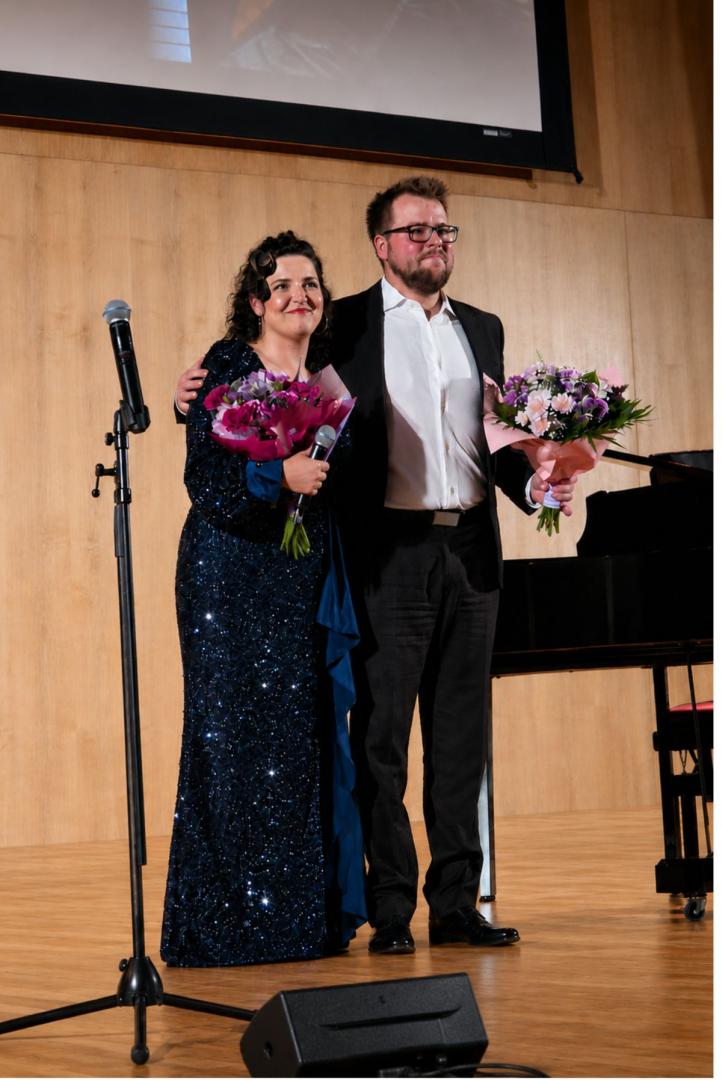 Wokalistka i pianista po koncercie stoją na scenie z bukietami kwiatów, obok mikrofonu i fortepianu.