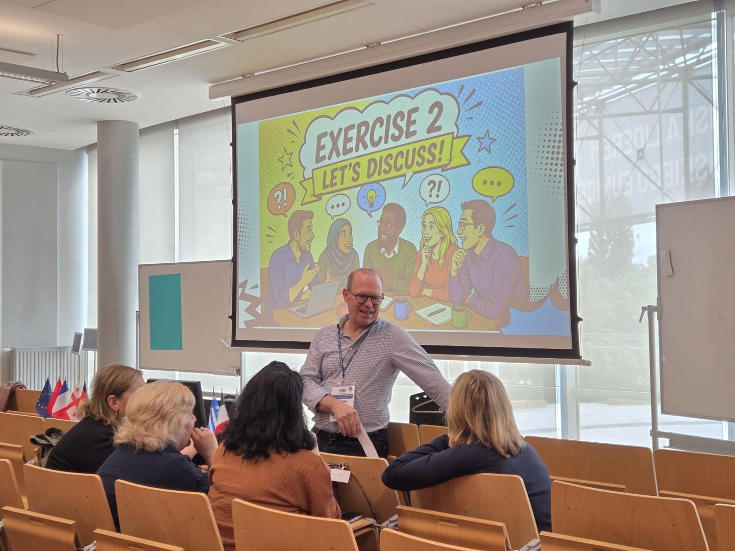 A speaker talking to workshop participants against a screen with the inscription "EXERCISE 2 LET'S DISCUSS!".