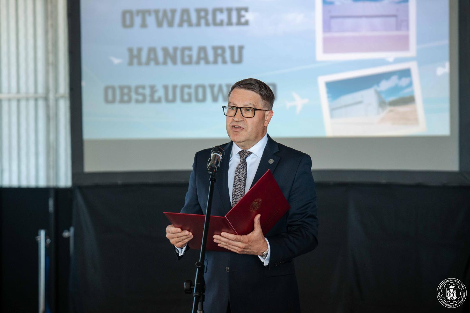 Rektor przemawia przy mikrofonie na tle ekranu z napisem „Otwarcie hangaru obsługowego”.