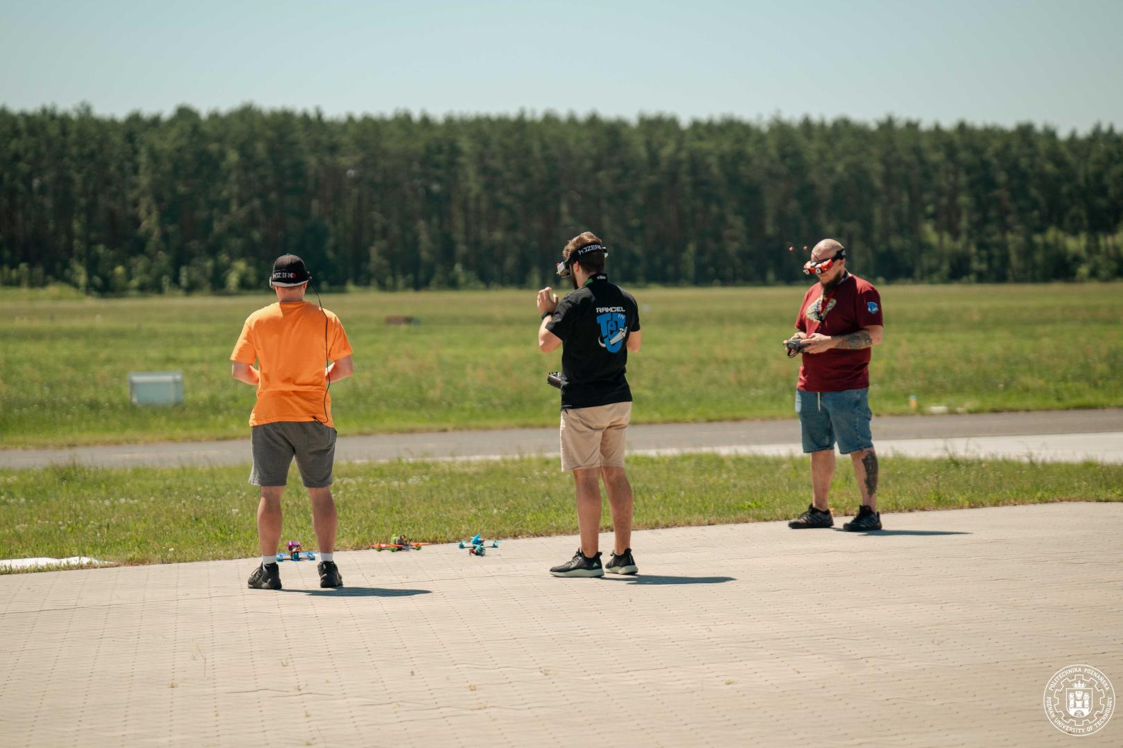 Trzej operatorzy w goglach FPV i z kontrolerami stoją przy dronach wyścigowych na płycie lotniska.