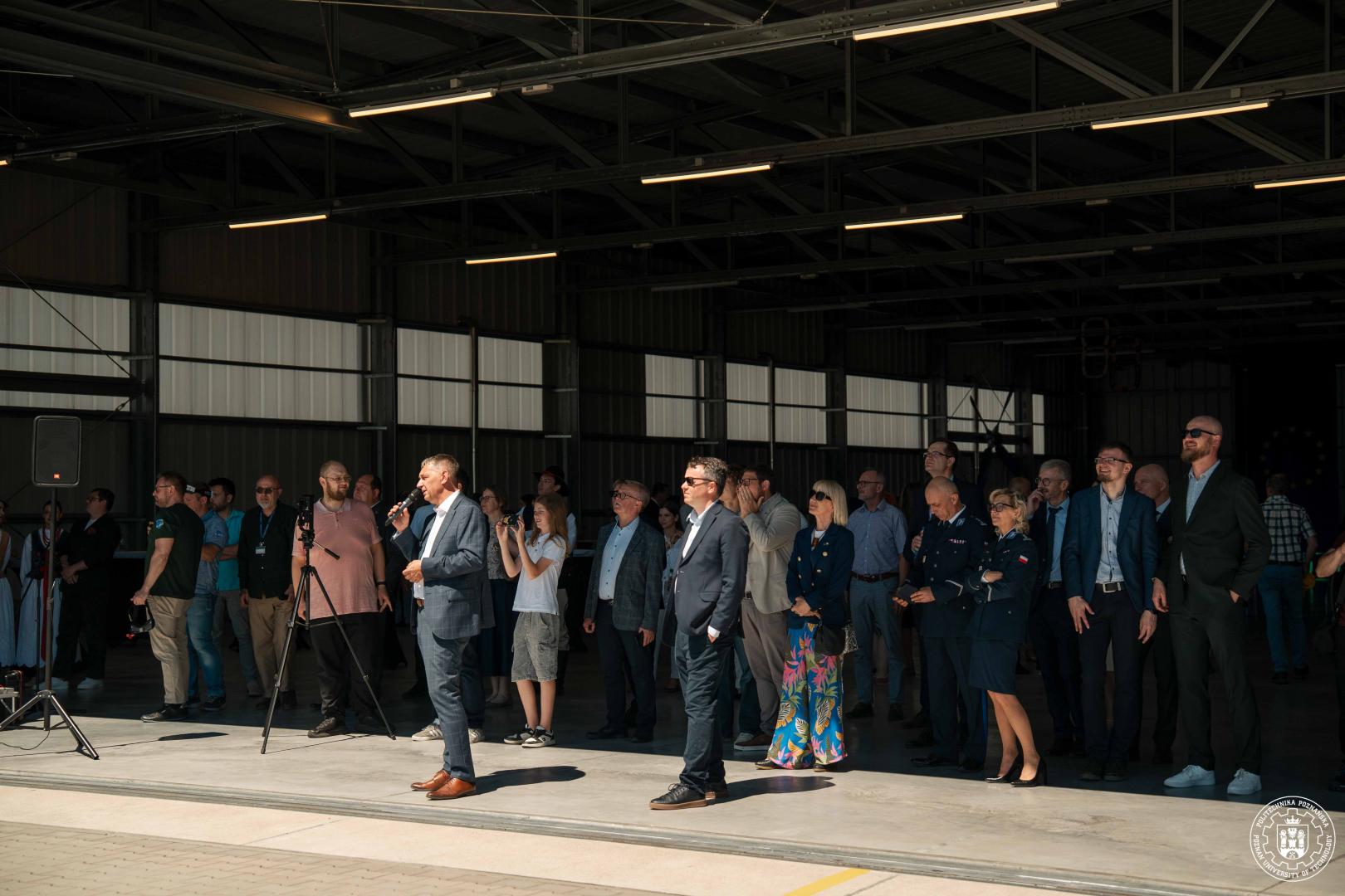Zgromadzeni wewnątrz hangaru goście słuchają przemówienia mężczyzny stojącego z mikrofonem po lewej stronie.