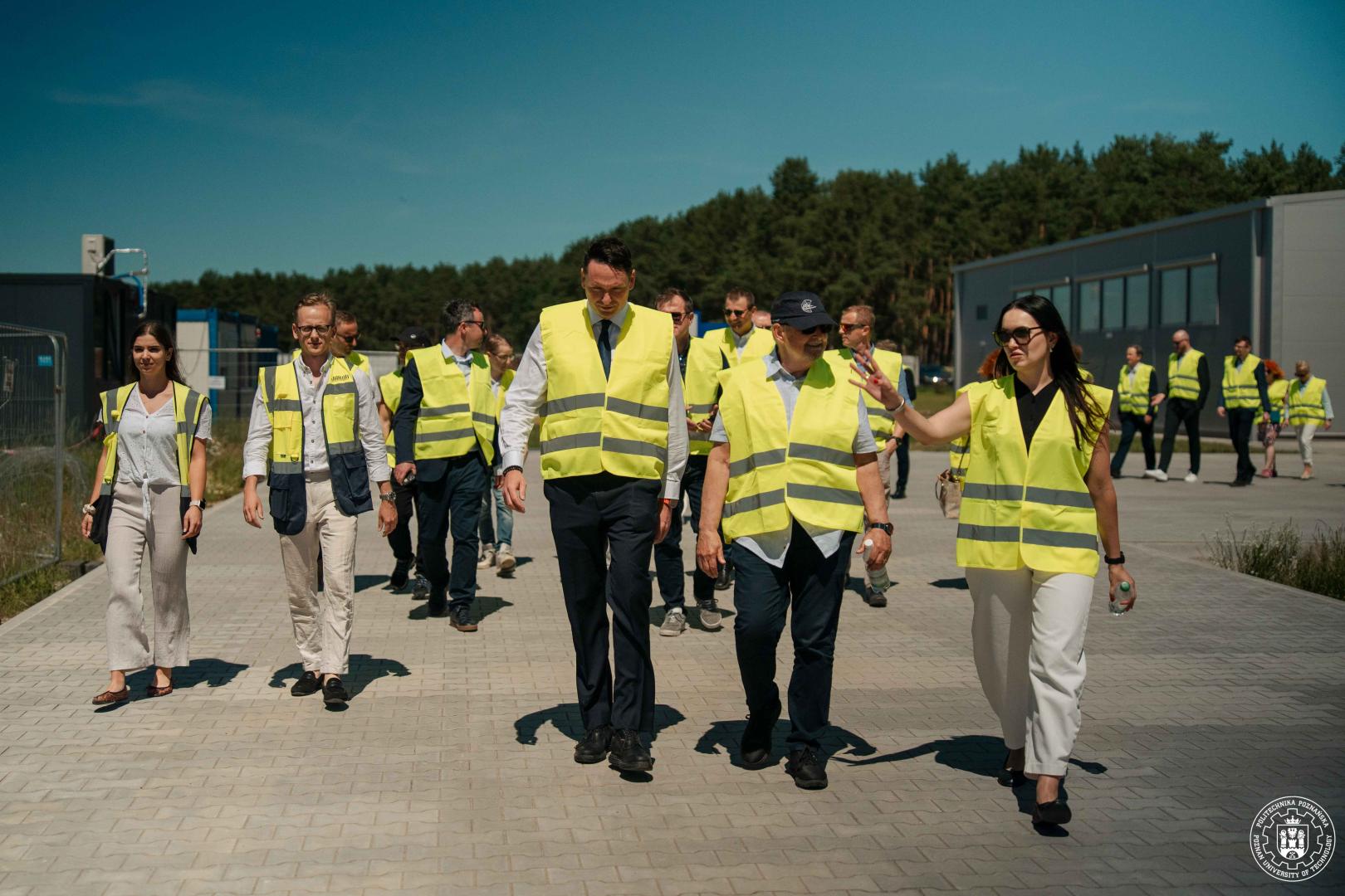 Grupa osób w odblaskowych kamizelkach spaceruje po brukowanej ścieżce na otwartym terenie.