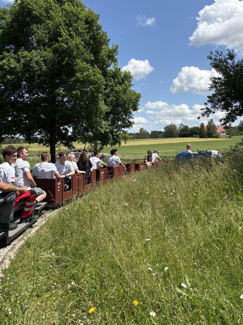 Miniaturowa kolejka zespołu PUTrain z pasażerami jadąca przez zielone, trawiaste tereny.