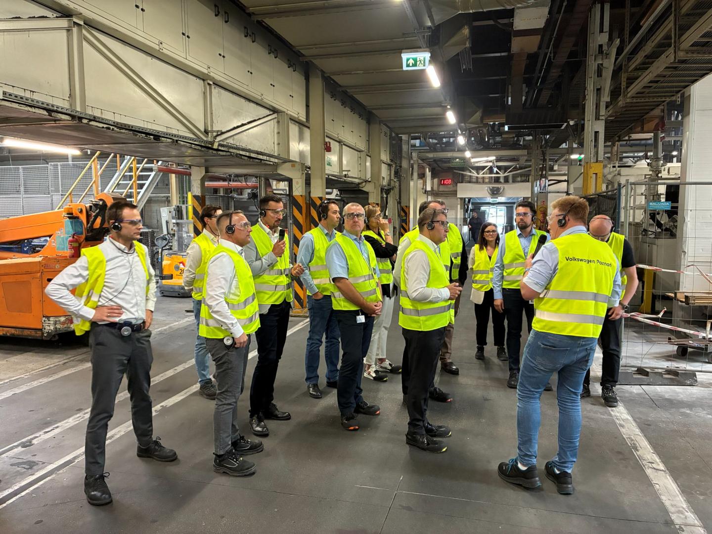 Delegacja w kamizelkach odblaskowych i słuchawkach słucha przewodnika podczas zwiedzania Odlewni Volkswagen Poznań.