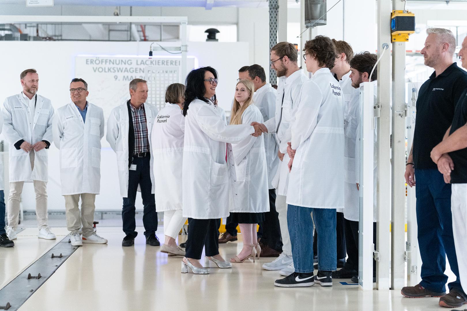 Przedstawiciele zarządu Volkswagen Poznań oraz studenci koła PUT Renovation w białych fartuchach laboratoryjnych podają sobie ręce podczas uroczystości w lakierni.