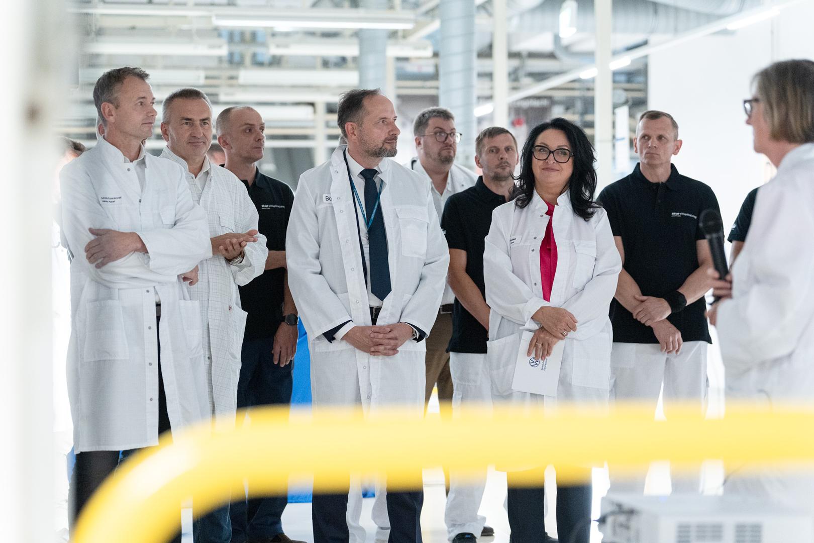 Zarząd Volkswagen Poznań oraz władze Politechniki Poznańskiej w białych fartuchach słuchają przemówienia podczas uroczystości w lakierni.