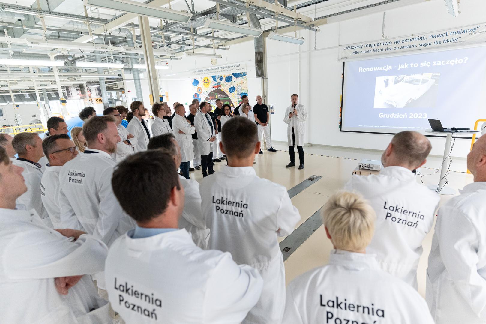 Zarząd Volkswagen Poznań oraz władze Politechniki Poznańskiej w białych fartuchach słuchają przemówienia podczas uroczystości w lakierni.