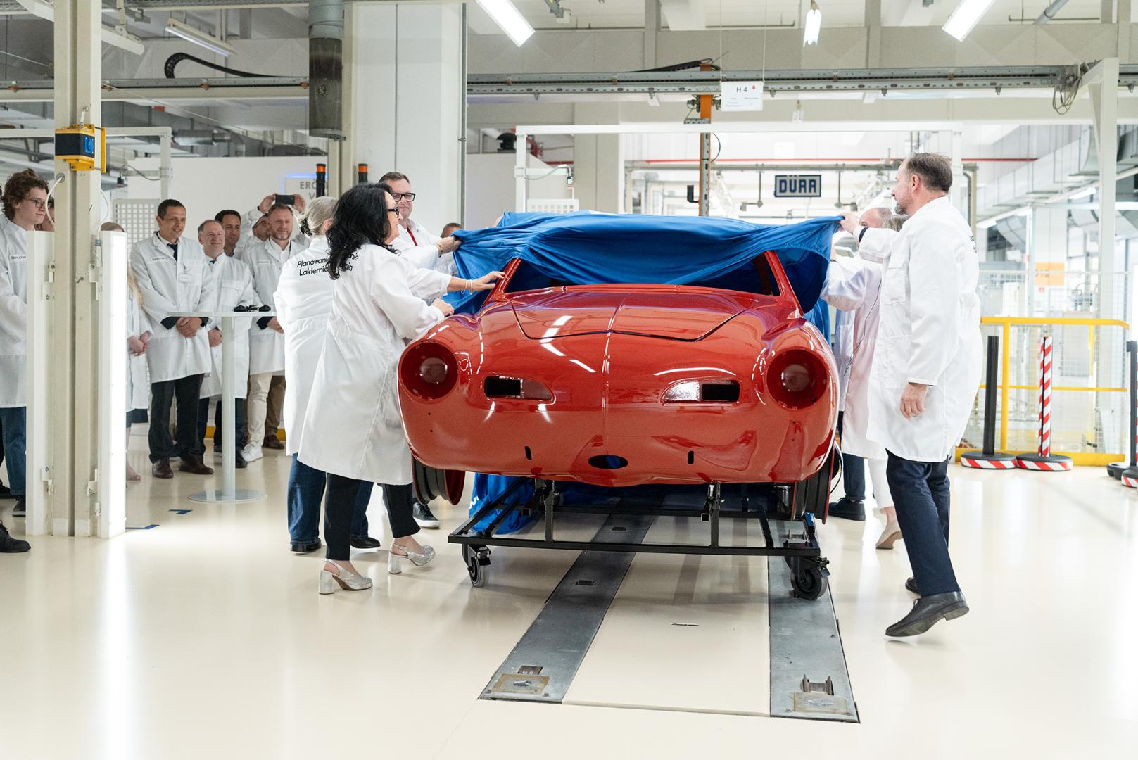 Oficjalne odsłonięcie odrestaurowanej, czerwonej karoserii Volkswagena Karmann Ghia poprzez ściągnięcie z niej niebieskiej płachty przez osoby w białych fartuchach.