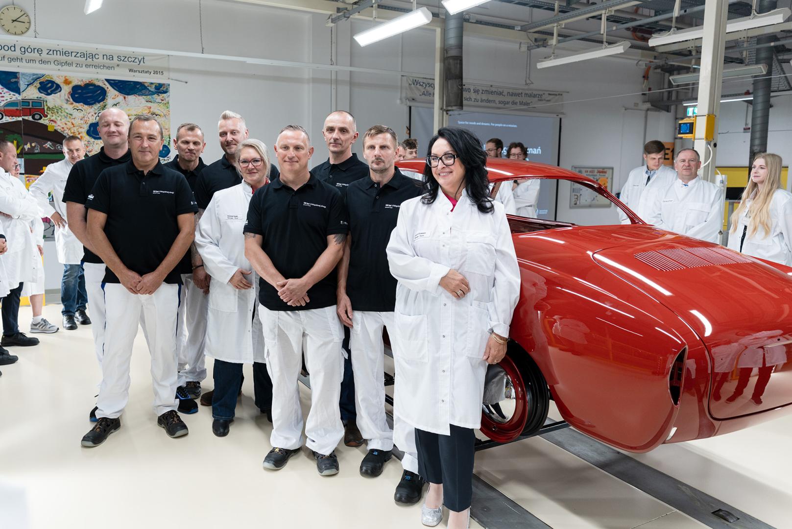 Pracownicy fabryki pozują wokół odrestaurowanego czerwonego Volkswagena Karmann Ghia w hali produkcyjnej.