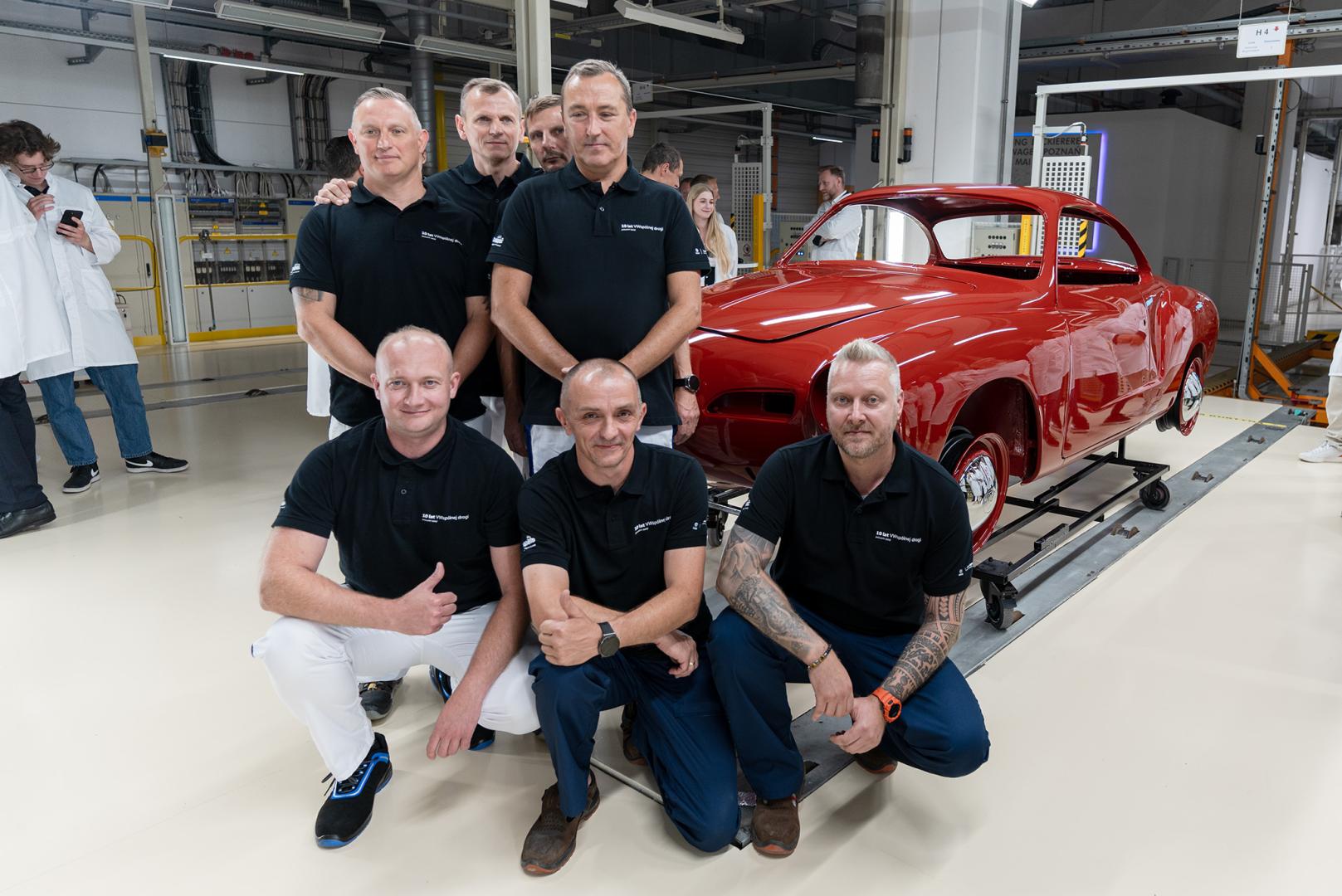 Pracownicy fabryki pozują wokół odrestaurowanego czerwonego Volkswagena Karmann Ghia w hali produkcyjnej.