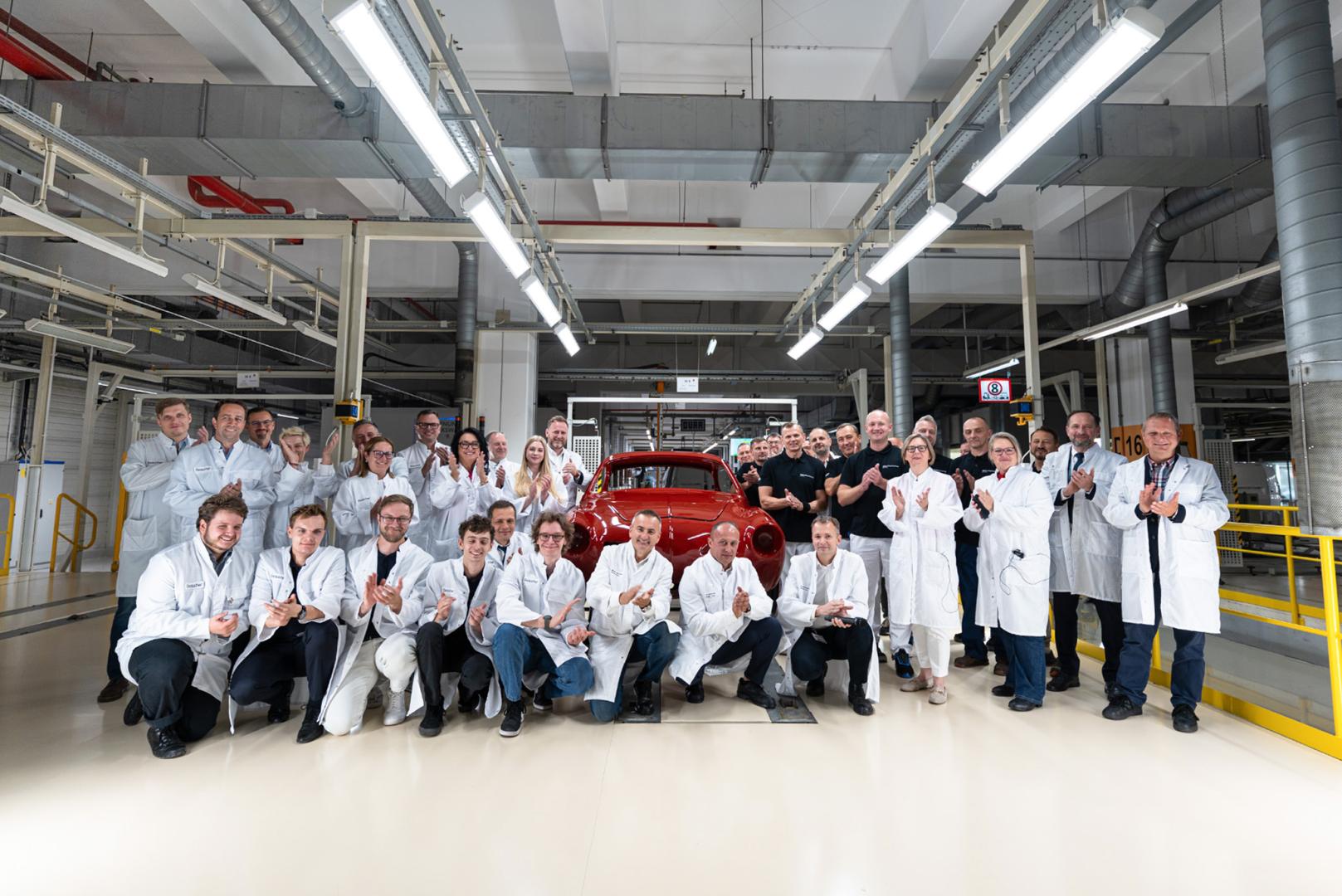 Pracownicy fabryki pozują wokół odrestaurowanego czerwonego Volkswagena Karmann Ghia w hali produkcyjnej.