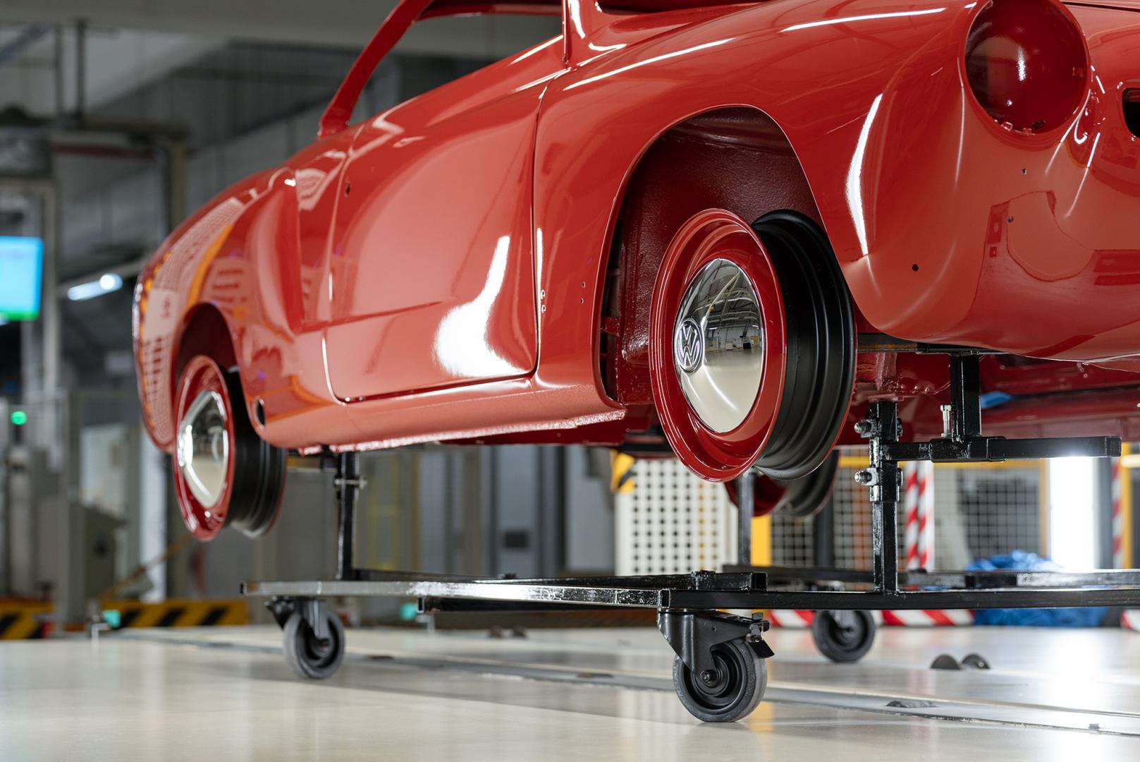 Błyszcząca, czerwona karoseria Volkswagena Karmann Ghia na wózku technologicznym, z widoczną czerwoną felgą i chromowanym kołpakiem z logo VW.
