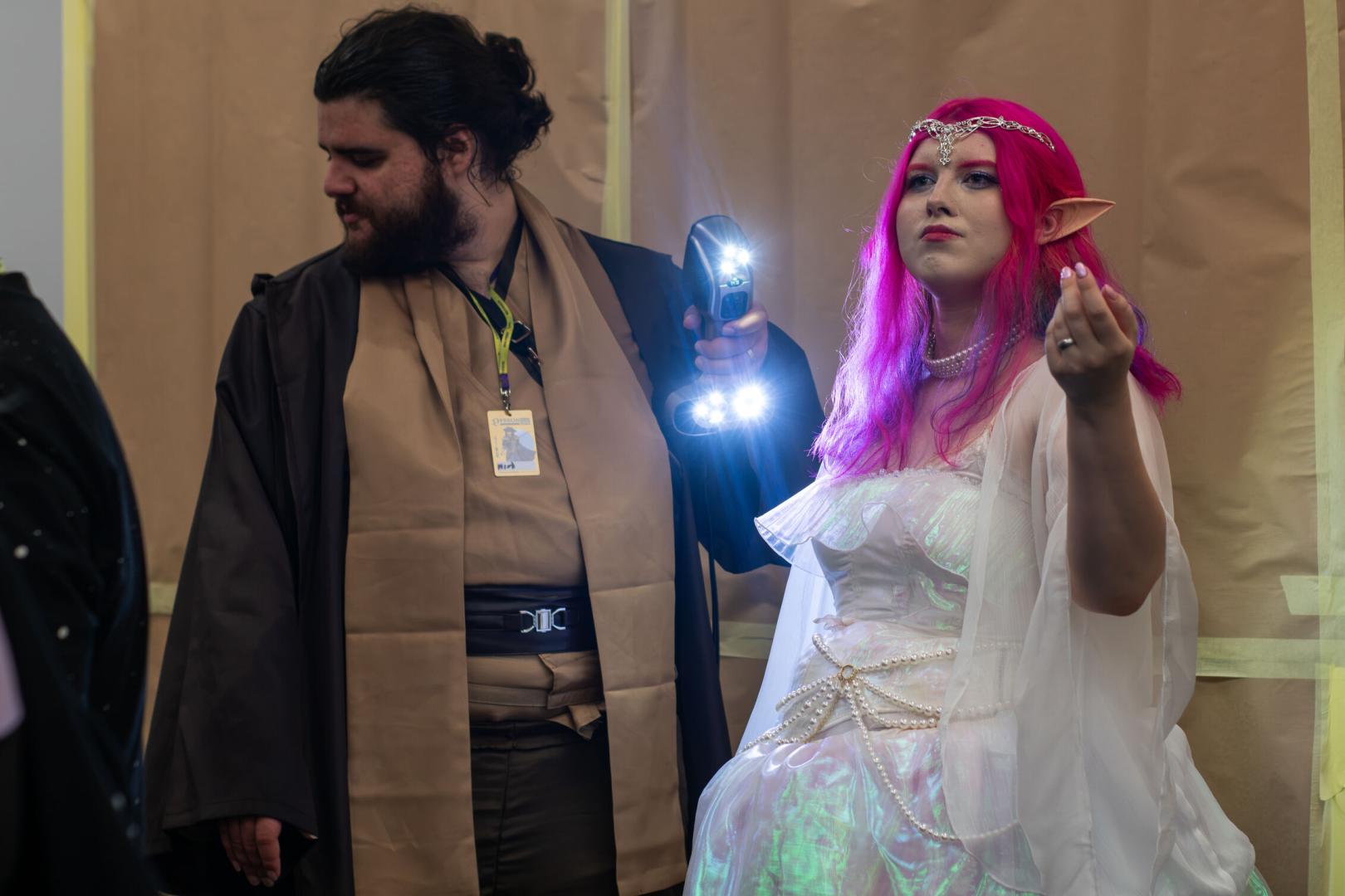 Dwoje cosplayerów: mężczyzna w brązowym płaszczu i kobieta w białej sukience z różowymi włosami oraz elfimi uszami, trzymająca świecącą latarkę.