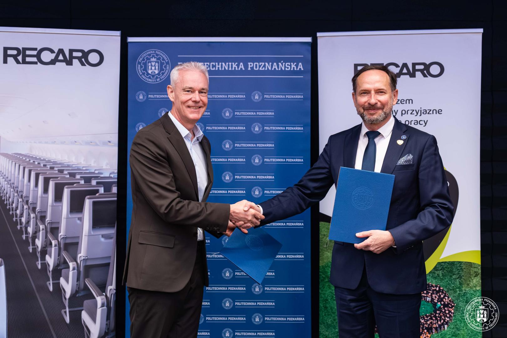 Prorektor dr hab. Arkadiusz Ptak, prof. PP i przedstawiciel firmy RECARO podają sobie dłonie z podpisanymi umowami na tle banerów Politechniki Poznańskiej.