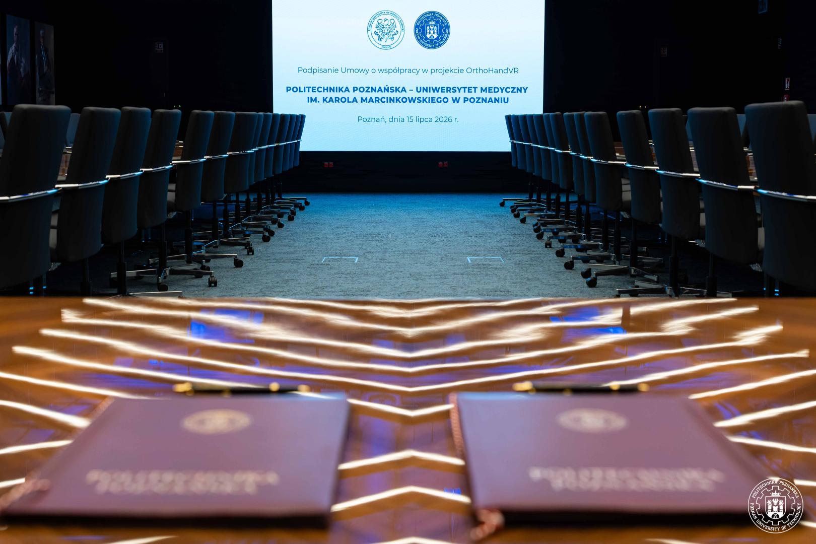Sala konferencyjna z rzędami krzeseł, stołem z dokumentami i ekranem wyświetlającym tytuł projektu OrthoHandVR oraz loga uczelni.