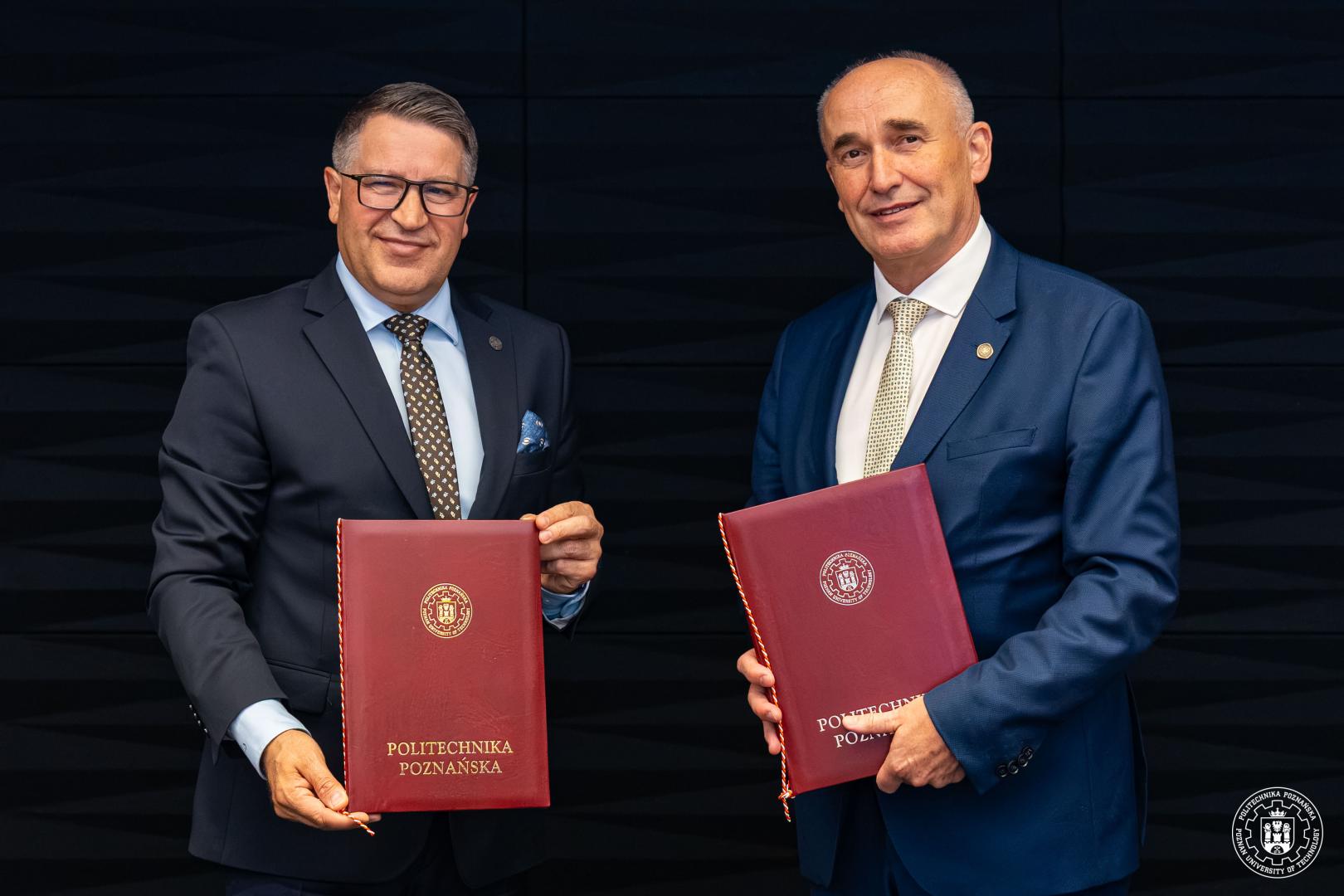 Dwaj mężczyźni w garniturach trzymają bordowe teczki z logo Politechniki Poznańskiej.