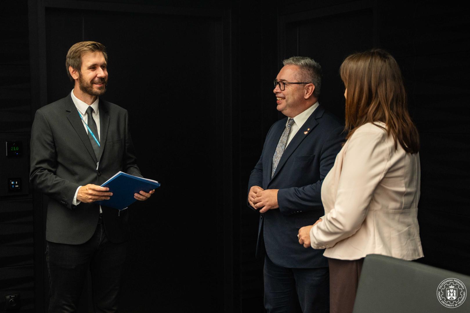 Trzy osoby w garniturach rozmawiają w Sali Senatu Politechniki Poznańskiej.