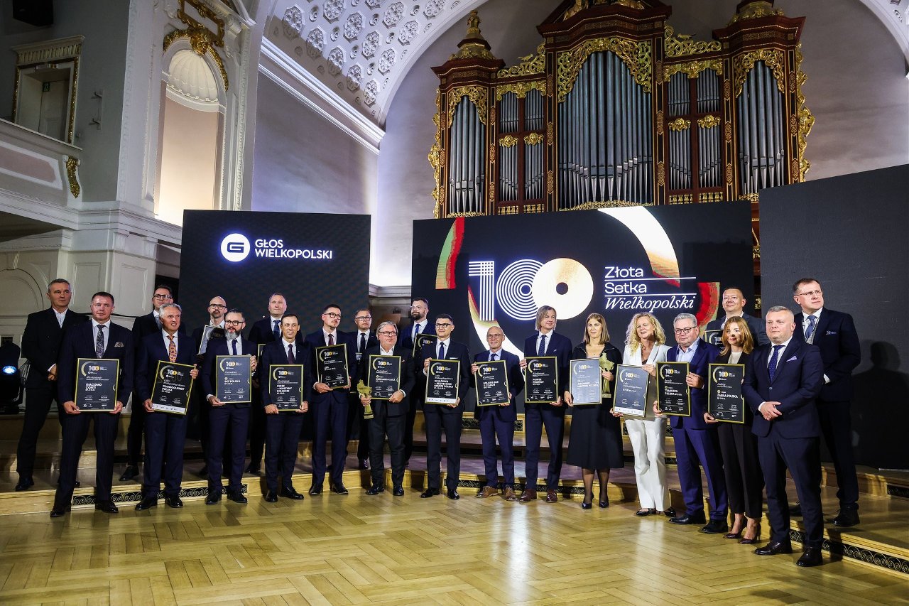Grupa około 20 osób w dwóch rzędach trzyma dyplomy ze złotymi ramkami na tle ekranu z napisem "Złota Setka Wielkopolski" i banera "GŁOS WIELKOPOLSKI" w auli uniwersyteckiej z organową fasadą.