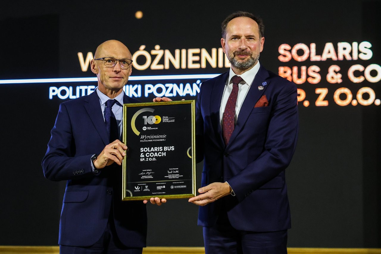 Dwaj mężczyźni stoją obok siebie, trzymając wspólnie oprawioną tablicę z wyróżnieniem, na tle podświetlonego napisu "WYRÓŻNIENIE POLITECHNIKI POZNAŃSKIEJ SOLARIS BUS & COACH SP. Z O.O.".