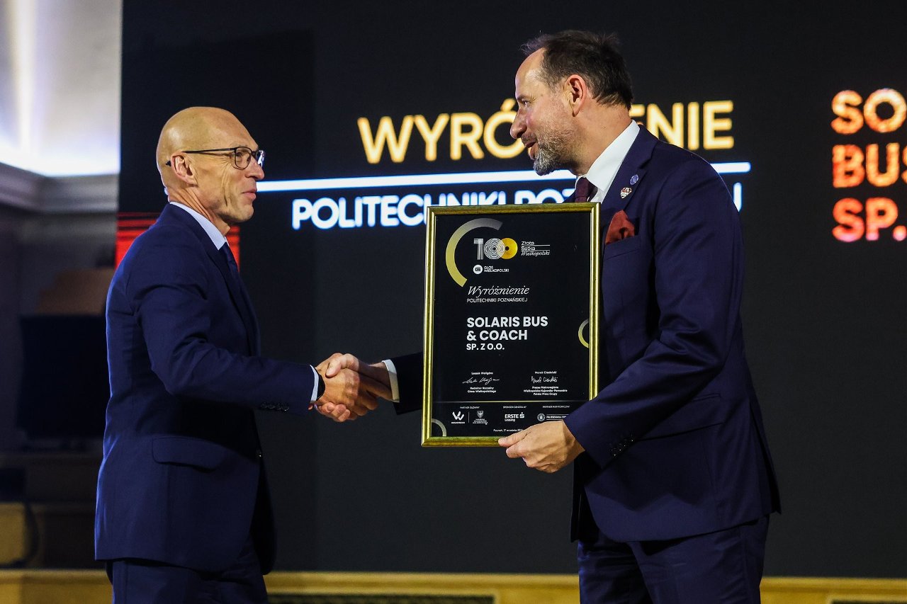 Dwaj mężczyźni w garniturach ściskają sobie dłonie: mężczyzna po lewej z okularami, po prawej z czerwonym krawatem i odznaką PP na klapie marynarki trzyma tablicę z napisem "Wyróżnienie POLITECHNIKI POZNAŃSKIEJ SOLARIS BUS & COACH SP. Z O.O.".