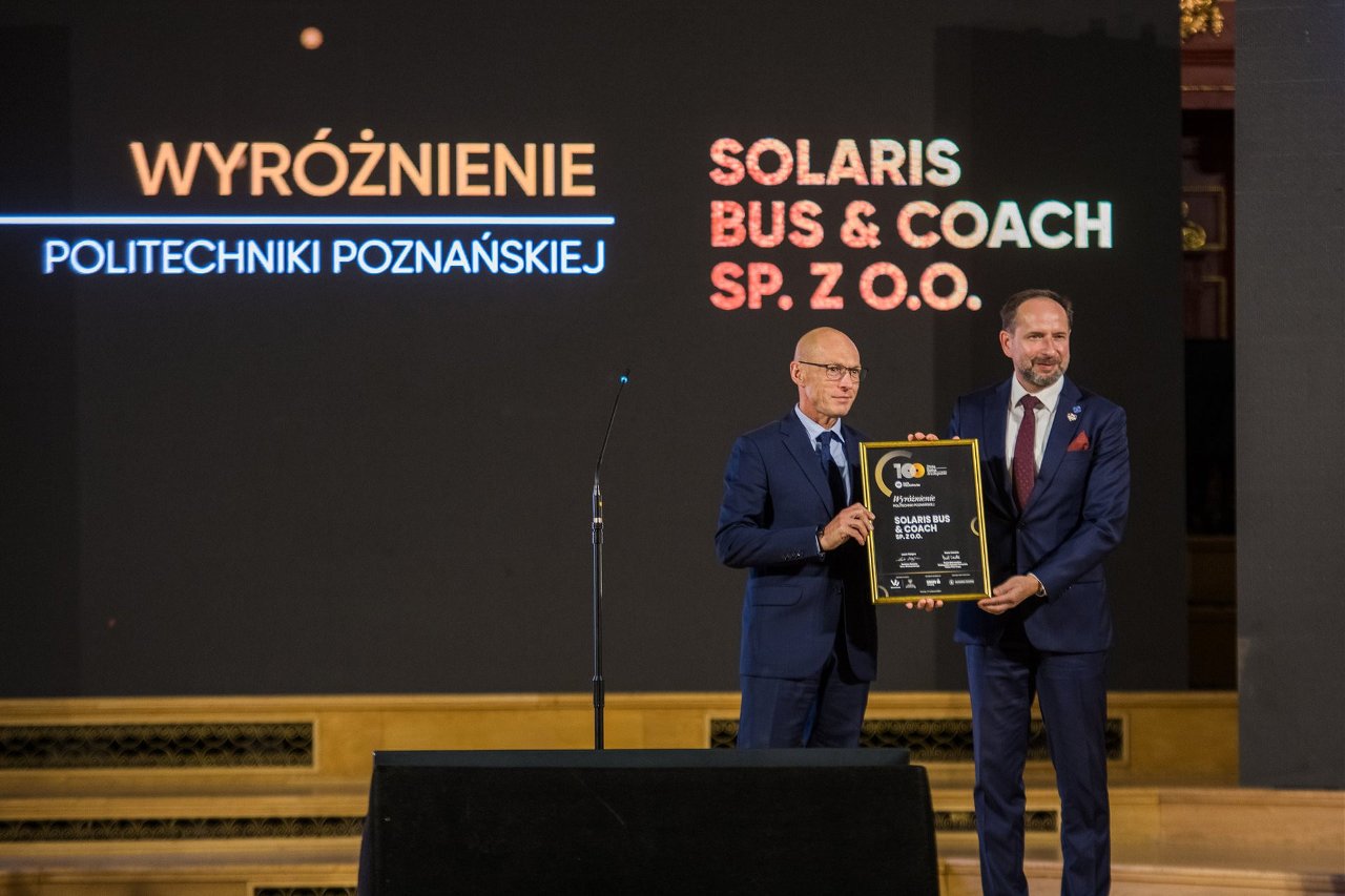 Dwaj mężczyźni na scenie podczas gali w ciemnych garniturach, trzymają w obu dłoniach oprawioną tablicę z wyróżnieniem "SOLARIS BUS & COACH SP. Z O.O.", a za nimi podświetlone napisy "WYRÓŻNIENIE POLITECHNIKI POZNAŃSKIEJ".
