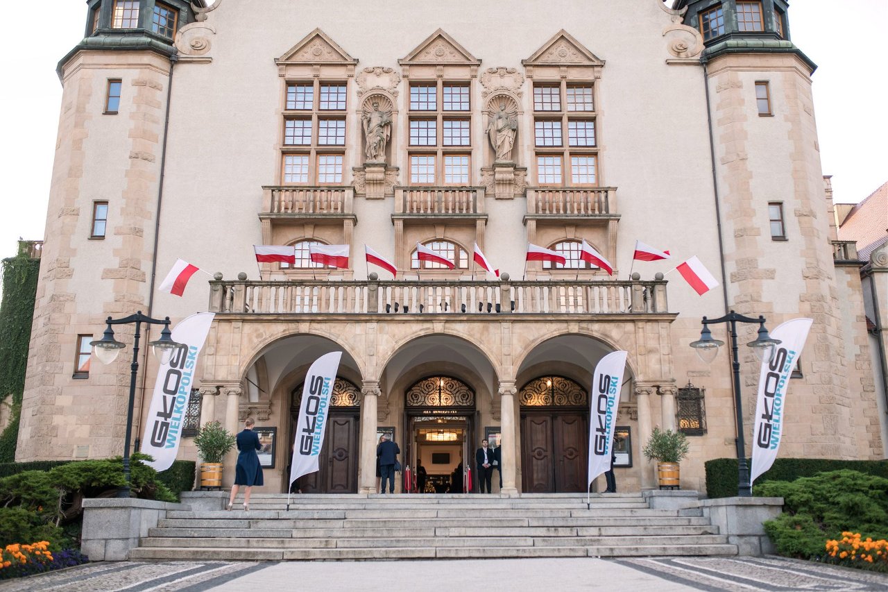 Fasada Auli Uniwersyteckiej z tabliczką "AULA UNIWERSYTECKA" przy wejściu, schodami i banerami "GŁOS WIELKOPOLSKI" po obu stronach, z flagami Polski i kilkoma osobami na schodach.