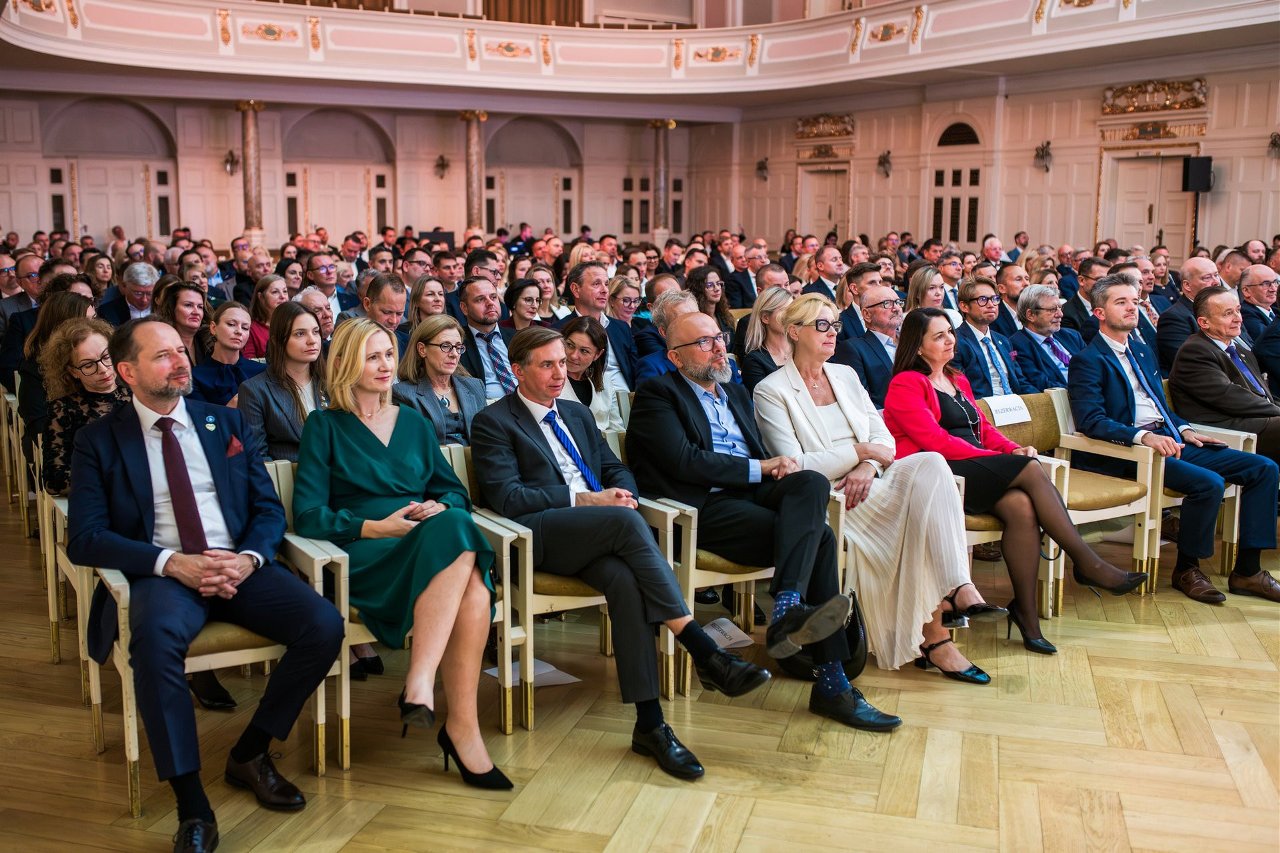 Sala konferencyjna podczas gali: około dwudziestu osób w eleganckich strojach siedzi w rzędach, zwróconych w stronę sceny, na tle bogato zdobionego wnętrza z drewnianą podłogą i złoceniami.