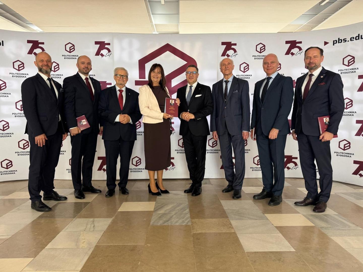 Ośmiu rektorów PP z prof. Jesionowskim po uroczystości nadania tytułu doktora honoris causa, na tle banera z napisem "Politechnika Bydgoska" i "75 lat". Prof. Misztal w brązowej sukni trzyma czerwoną oprawę z portretem uhonorowanego.