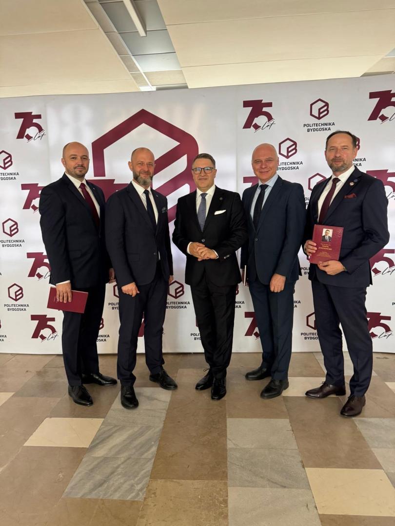 Pięciu rektorów PP w garniturach po uroczystości nadania tytułu doktora honoris causa, na tle banera z logo Politechniki Bydgoskiej i napisem "75 lat". Jeden z mężczyzn trzyma czerwoną okładkę z portretem uhonorowanego.