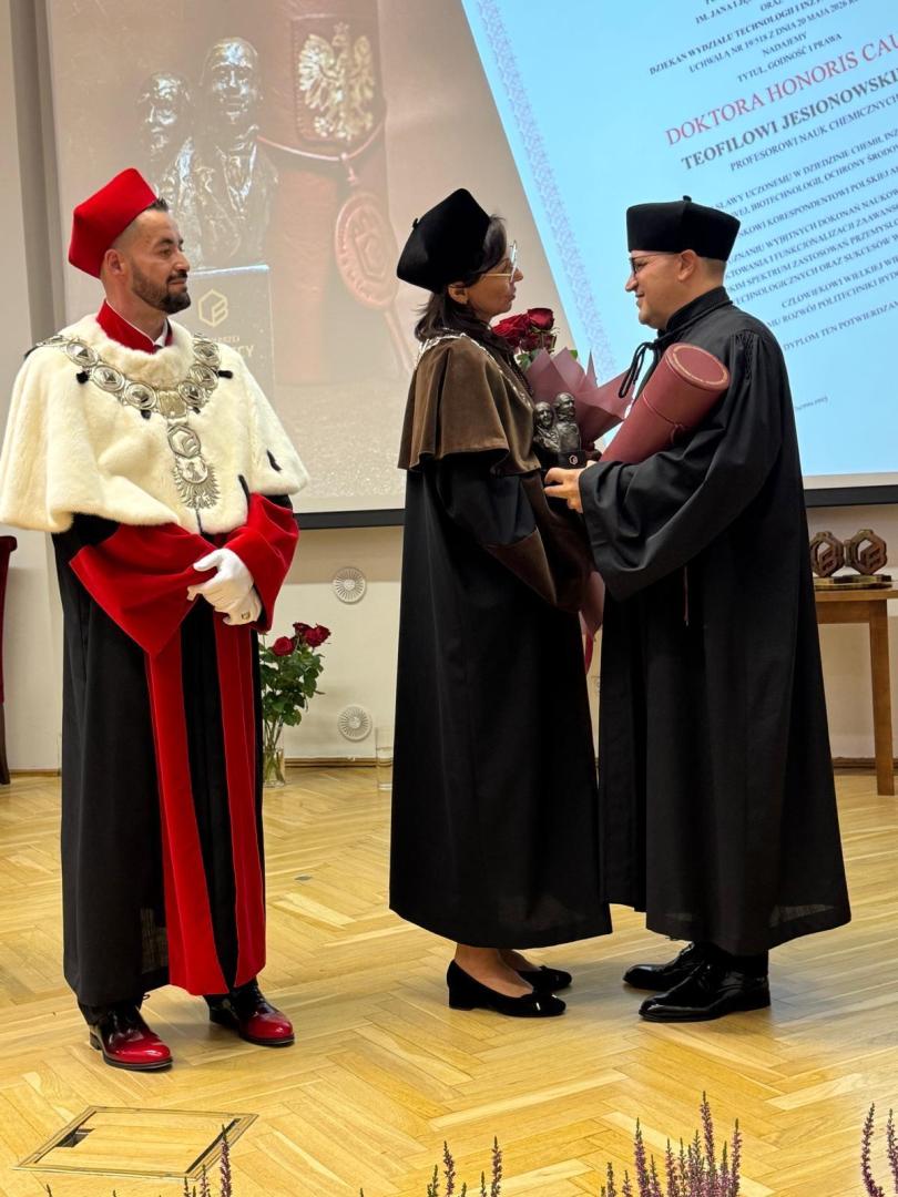 Uroczystość nadania tytułu doktora honoris causa w sali z drewnianą podłogą. Kobieta w todze trzyma bukiet róż i statuetkę, prof. Jesionowski w czarnej todze podaje jej dłoń, a w tle ekran z napisem "DOKTORA HONORIS CAUSA TEOFILA JESIONOWSKIEGO".