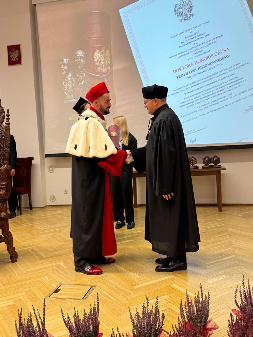 Dwóch mężczyzn w togach akademickich (prof. Jesionowski z prawej) ściska dłonie podczas ceremonii nadania tytułu doktora honoris causa. Mężczyzna po lewej w czerwonej todze z białym futrem trzyma upominek z herbem RP, a na ścianie baner z napisem "DOKTORA HONORIS CAUSA TEOFILA JESIONOWSKIEMU".
