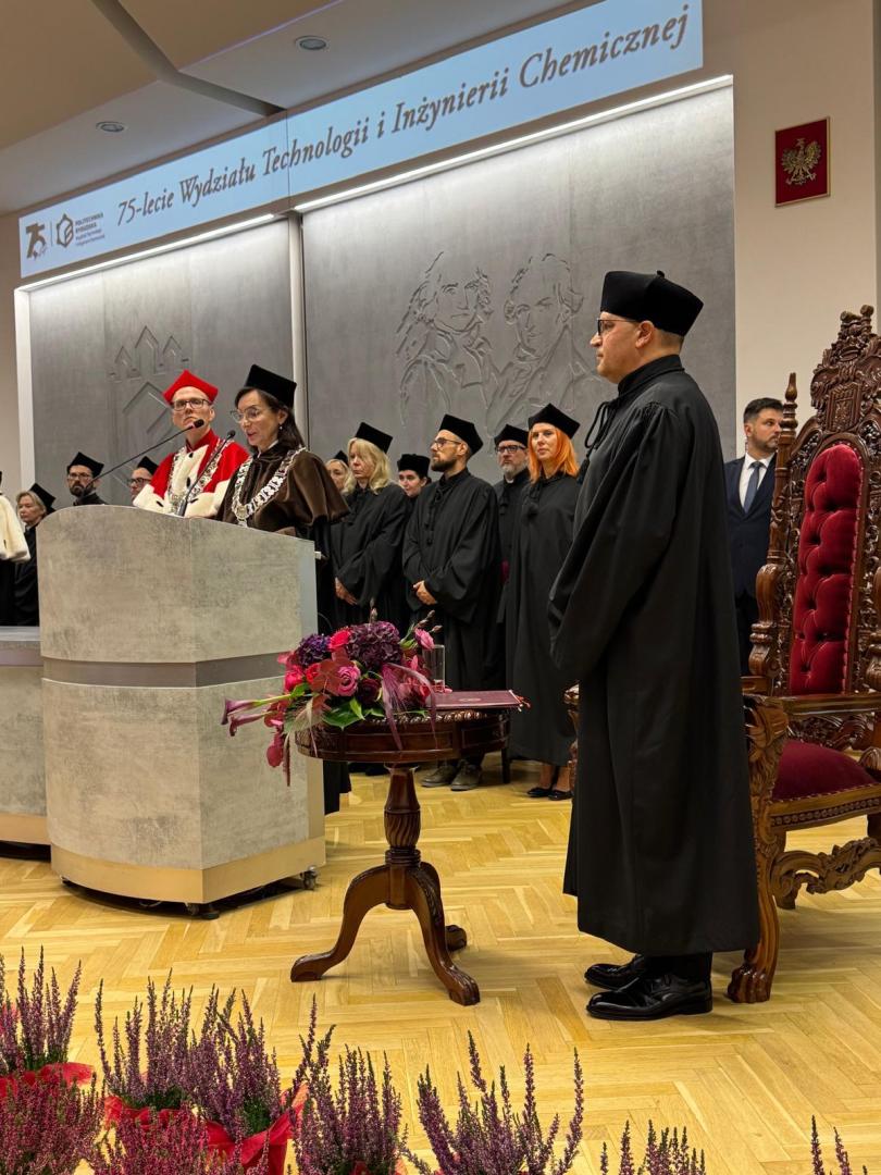 Ceremonia nadania tytułu doktora honoris causa w sali z banerem "75-lecie Wydziału Technologii i Inżynierii Chemicznej". Kobieta w czarnej todze przemawia przy pulpicie, w tle grupa osób w togach akademickich. Po prawej stronie pulpitu stoi prof. Jesionowski w czarnej todze akademickiej i czarnym berecie, zwrócony w stronę pulpitu. Za nim ozdobny, drewniany fotel z czerwonym obiciem.