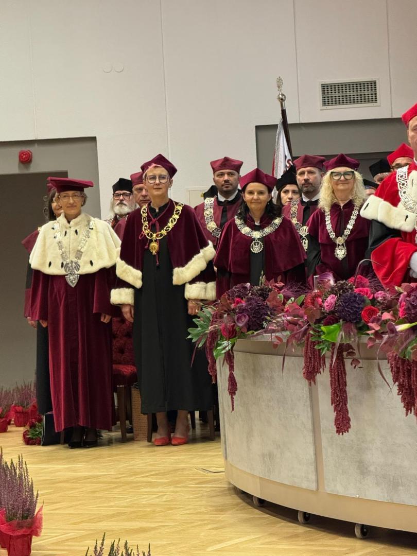 Grupa rektorów i dziekanów w bordowych togach z białymi obszyciami i medalami, a w tle flaga Polski i dekoracja kwiatowa na okrągłym stole.