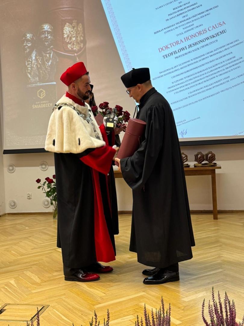 Dwóch mężczyzn w togach akademickich podczas aktu wręczenia tytułu doktora honoris causa. Mężczyzna w czerwonej todze z białym futrem wręcza bukiet róż i pojemnik z wstęgą prof. Jesionowskiemu.