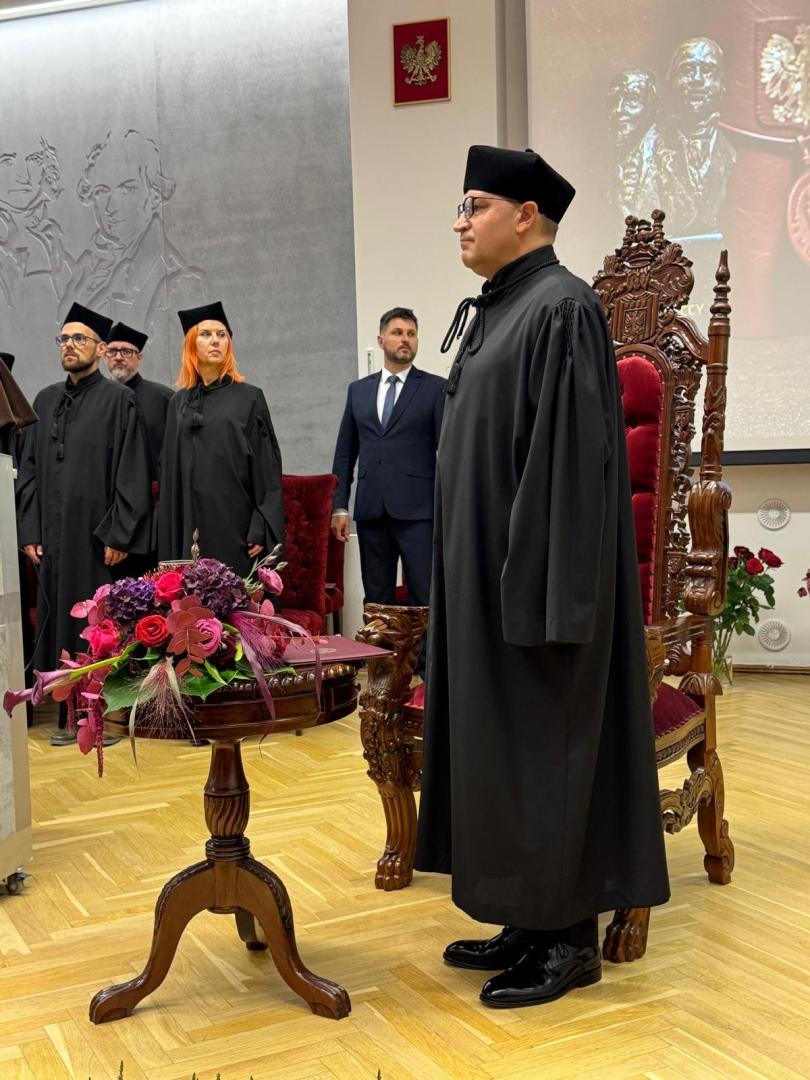 Prof. Jesionowski w czarnej todze akademickiej stoi na scenie w sali z ceremonialnym fotelem i tablicą pamiątkową. Na stoliku po lewej bukiet różowych i fioletowych kwiatów, a na ścianie herb z orłem.
