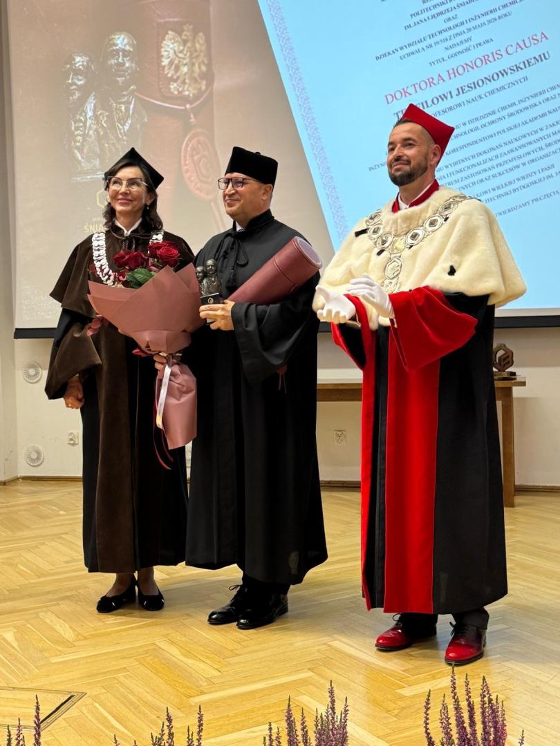 Uroczystość nadania tytułu doktora honoris causa. Kobieta w brązowej todze trzyma bukiet róż, mężczyzna w czarnej todze wręcza zwinięty dyplom, a w tle ekran z treścią oświadczenia nadania tytułu.