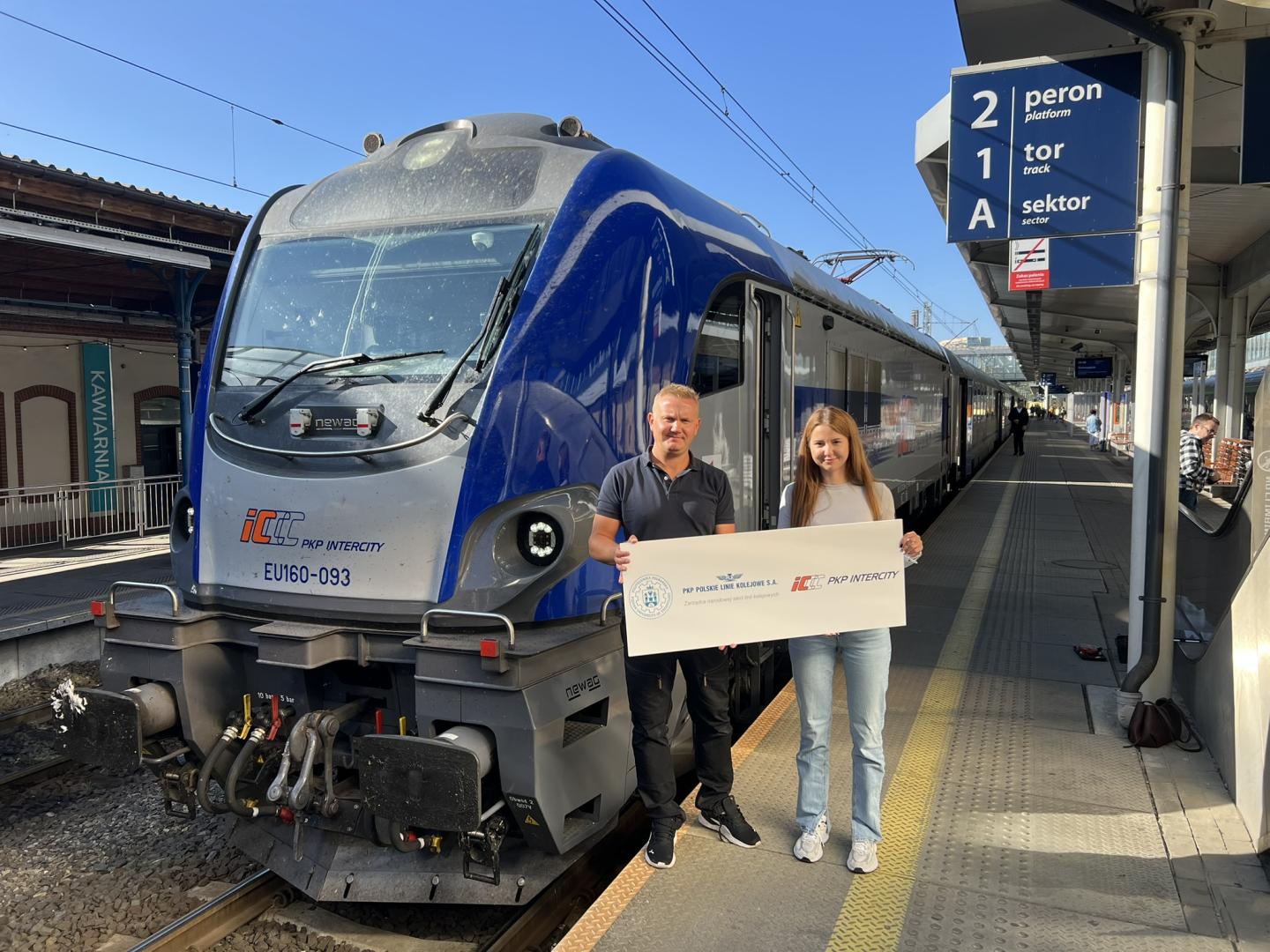 Przedstawiciele PKP Intercity i PKP PLK z planszą informacyjną przed pociągiem na stacji.