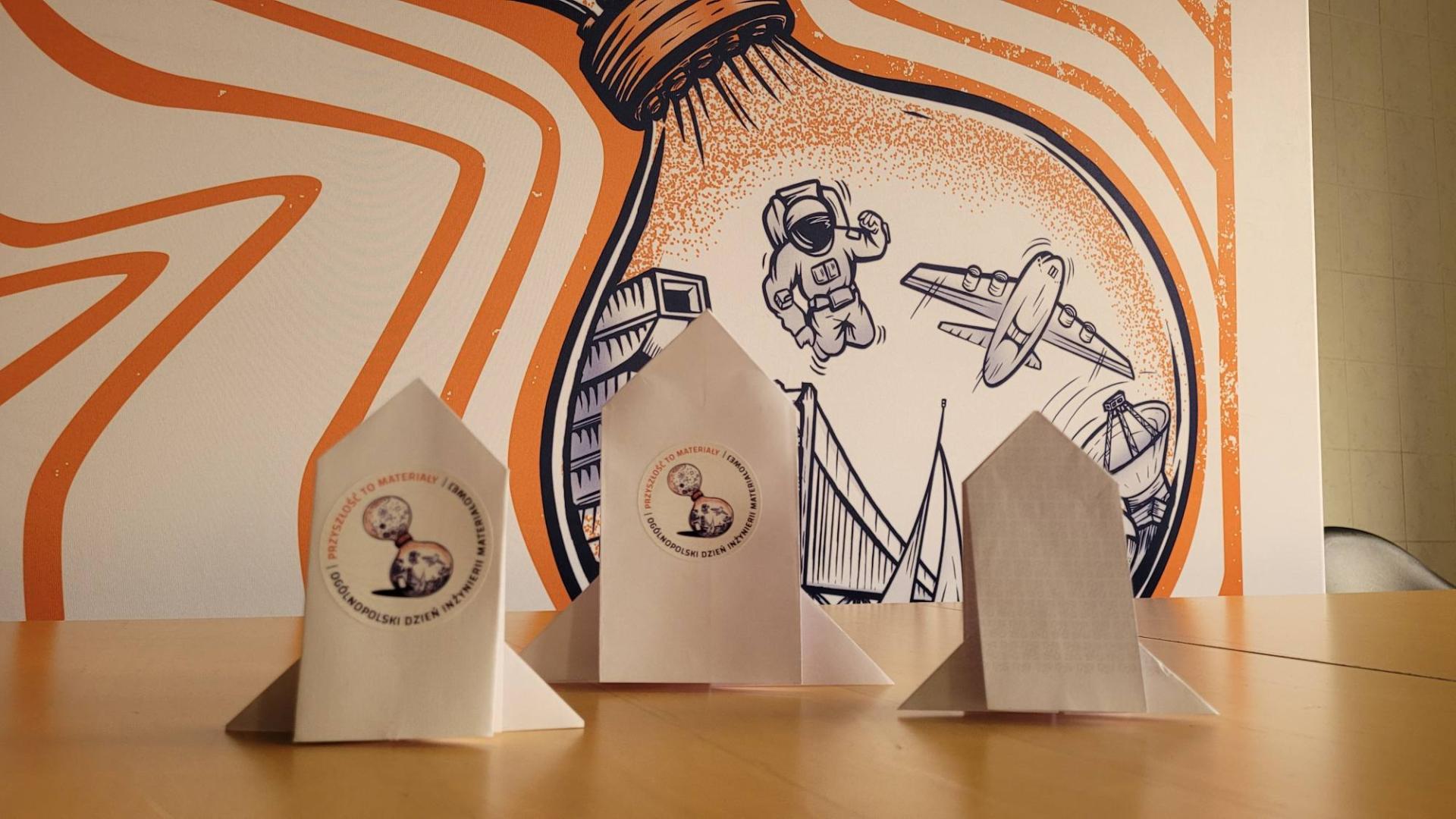 Trzy papierowe rakiety origami z logo Ogólnopolskiego Dnia Inżynierii Materiałowej na tle grafiki o tematyce kosmicznej.
