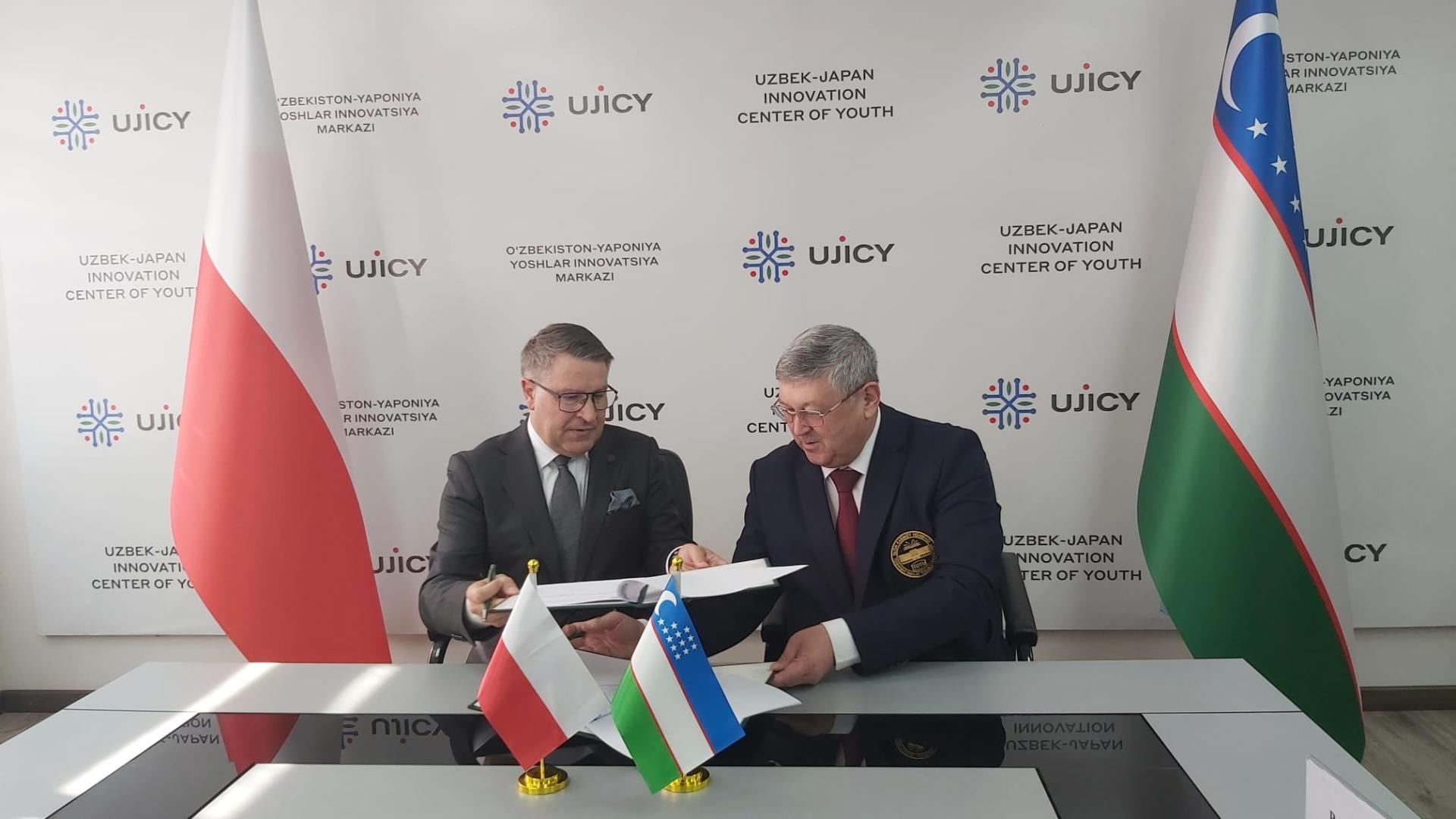 Dwaj mężczyźni wymieniają się dokumentami nad błyszczącym blatem stołu, w którym odbijają się ich sylwetki. Mężczyzna po prawej stronie przesuwa dokumenty po stole, a mężczyzna po lewej podaje je nad nimi.