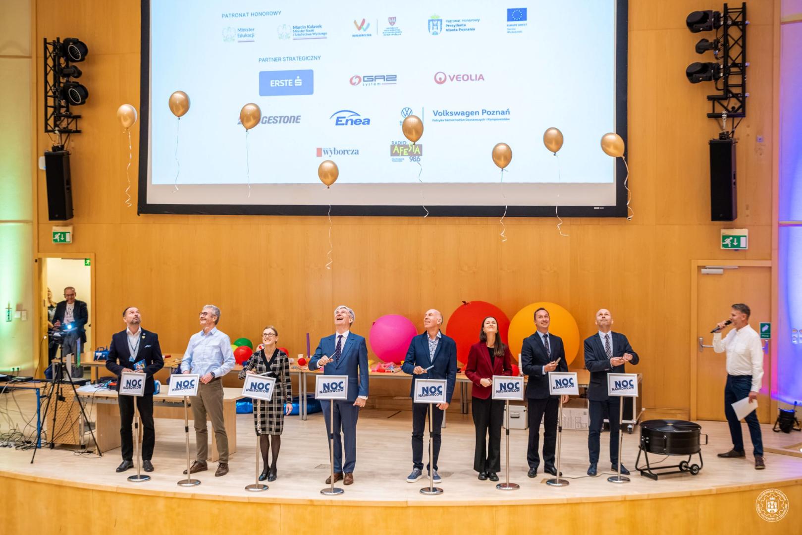 Organizatorzy na scenie Politechniki Poznańskiej z tabliczkami „NOC NAUKOWCÓW” patrzą na szybujące w górę złote balony.