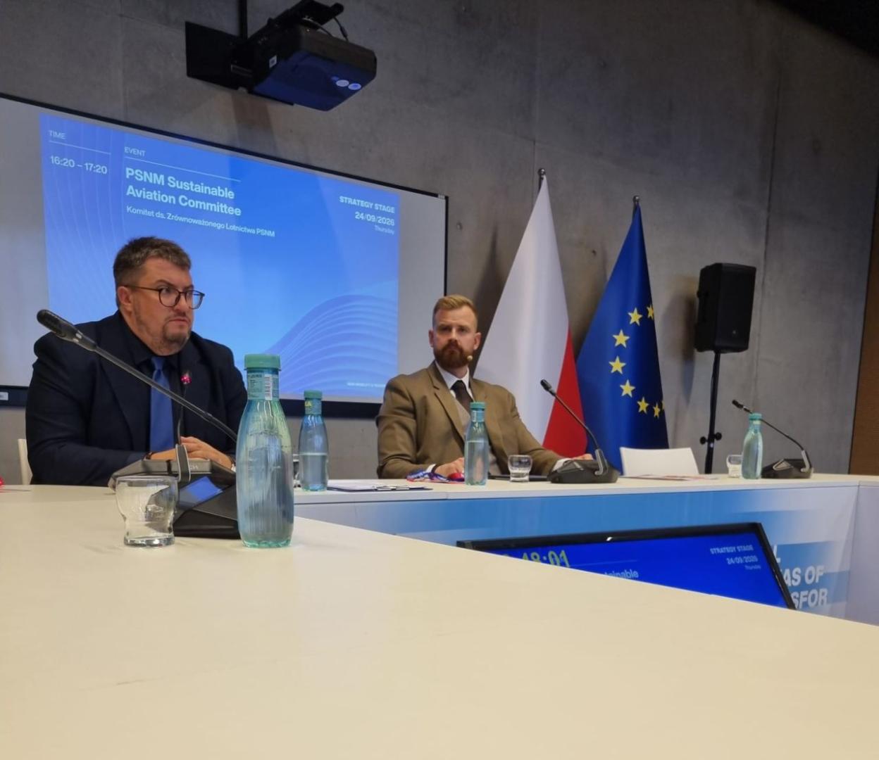Dwaj mężczyźni siedzą za długim, białym stołem konferencyjnym na tle flag Polski i Unii Europejskiej. Za nimi znajduje się ekran z napisem „PSNM Sustainable Aviation Committee” oraz „Komitet ds. Zrównoważonego Lotnictwa PSNM”.