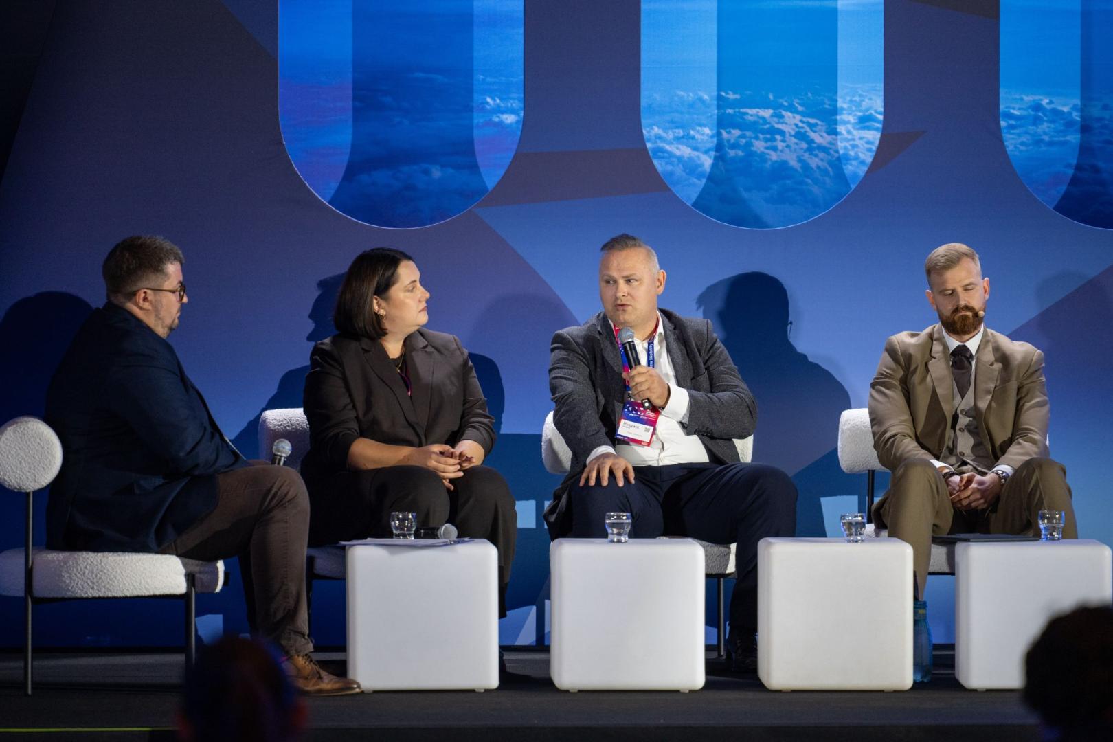 Panel dyskusyjny na scenie z udziałem czterech osób siedzących na tle niebieskiej ścianki z grafiką okien samolotu. Mężczyzna w centrum mówi do mikrofonu, a na jego identyfikatorze widnieje napis „Moderator”. Przed uczestnikami stoją podświetlane stoliki w kształcie sześcianów.