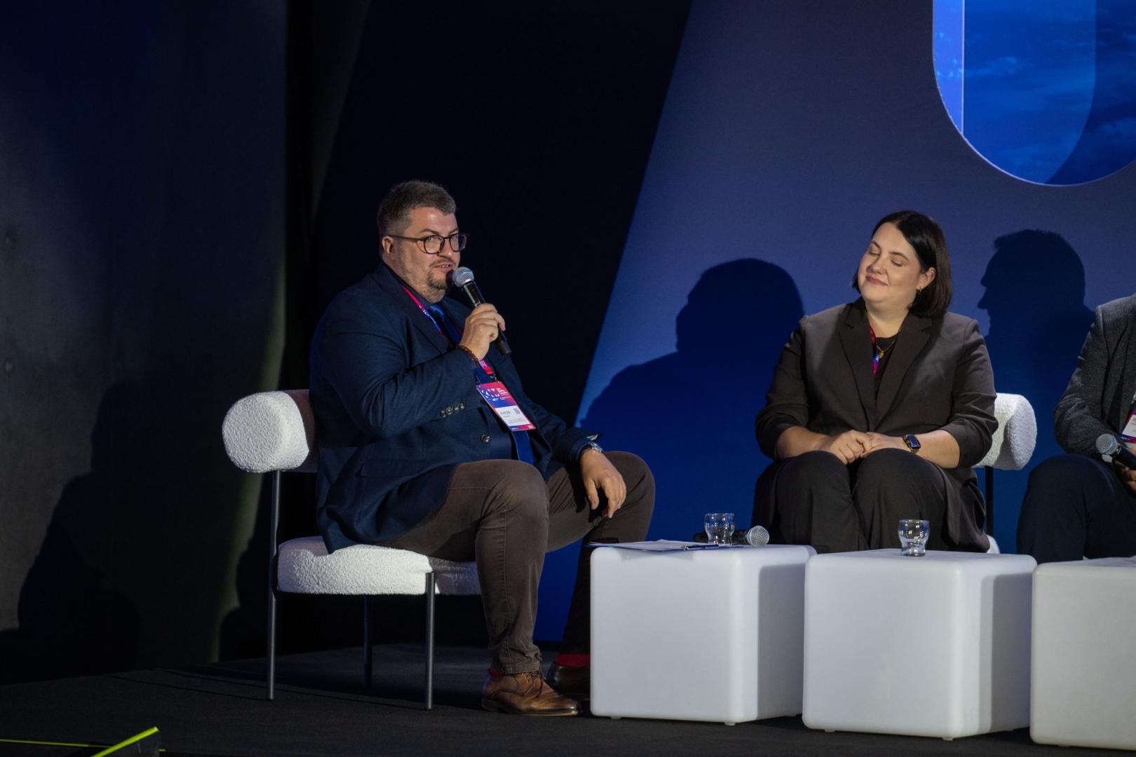 Mężczyzna w okularach i ciemnoniebieskiej marynarce mówi do mikrofonu podczas panelu dyskusyjnego. Obok niego siedzi kobieta w ciemnoszarym garniturze ze splecionymi dłońmi, patrzy w jego stronę. Na ich identyfikatorach widnieje różowo-niebieski napis „KNM”.