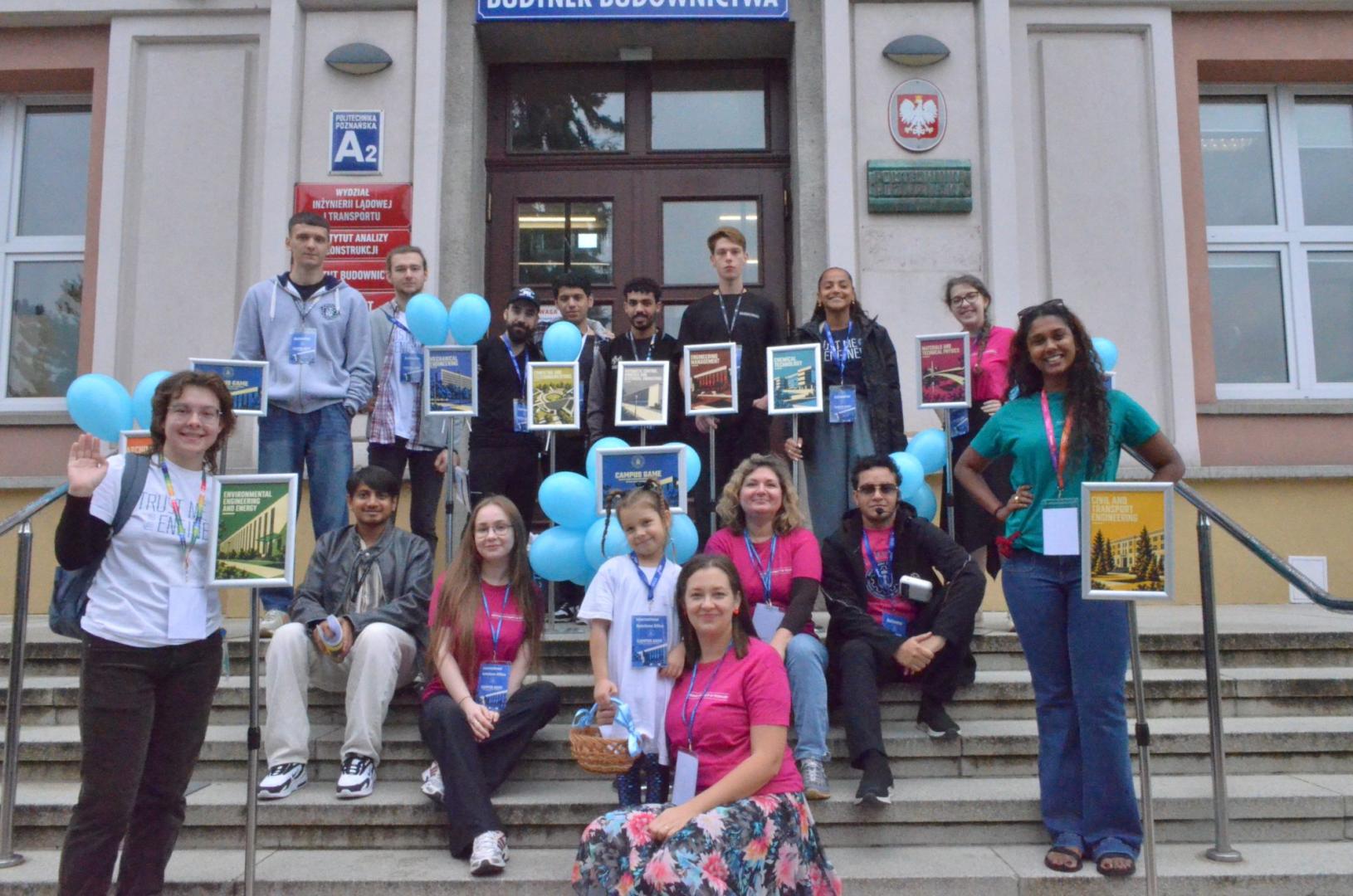 A group of international students and organizers poses for a photo on the stairs in front of the Building of Civil Engineering at Poznan University of Technology, holding blue balloons and signs with the names of the faculties.