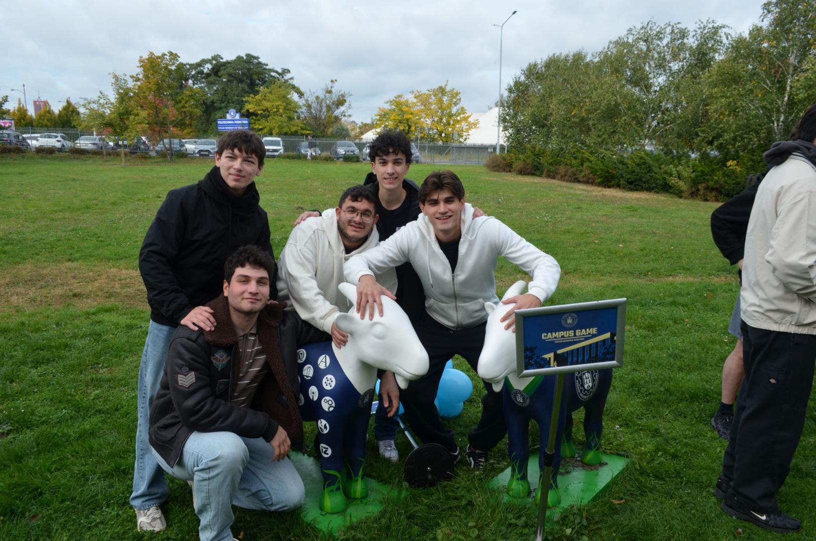 Five students pose with two goat sculptures on a lawn. A sign with the slogan "CAMPUS GAME" is visible next to one of them.