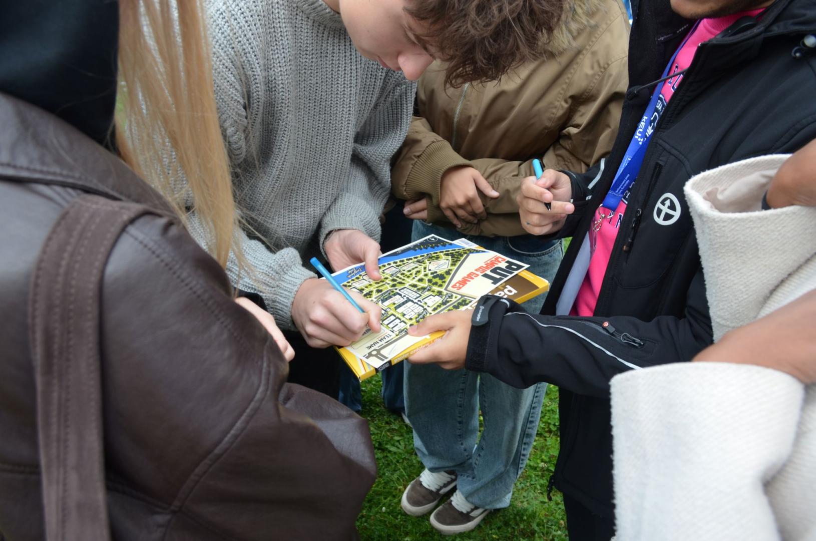 A group of students fills out a city game form with a campus map and the text "PUT CAMPUS GAME" using a pen.