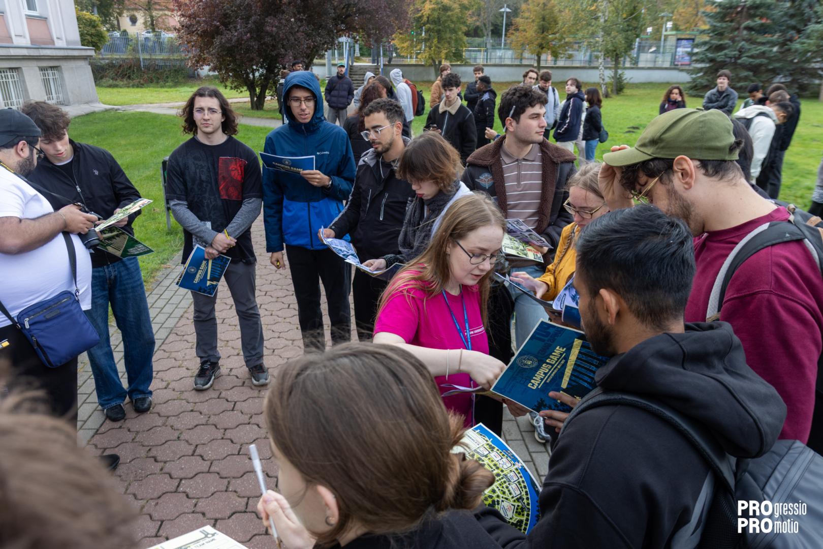 A group of students gathered outdoors analyzes materials with the text "Campus Game" and campus maps, assisted by an organizer in a pink shirt.