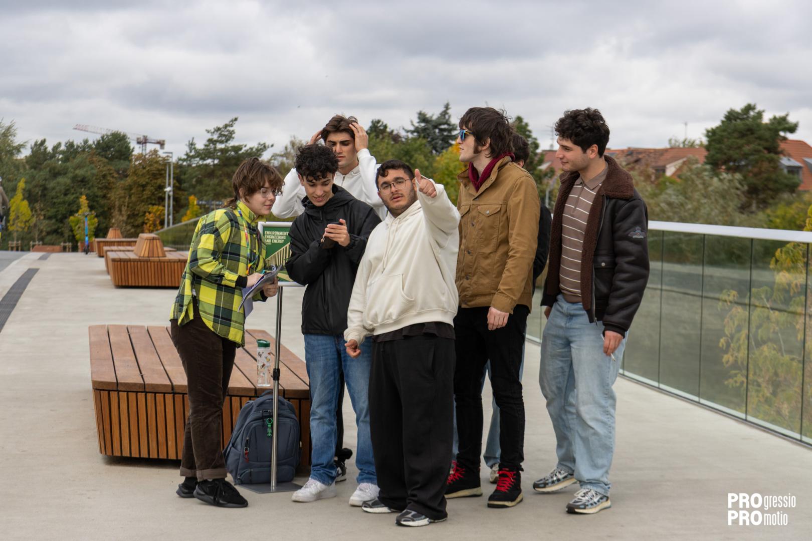 A group of students stands on a bridge or terrace, where one of them points a direction during a field task.